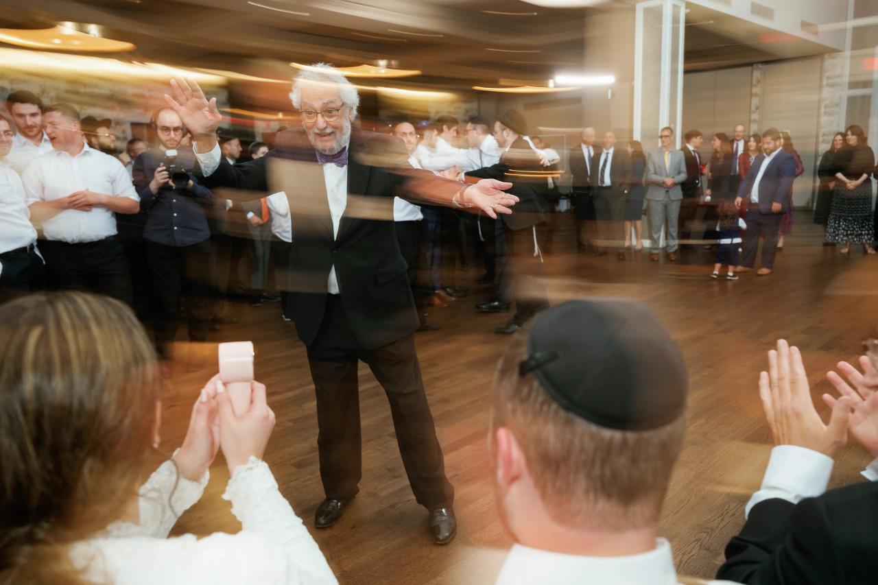 Indoor celebration dance-floor scene with a formally dressed man extending his hands while guests watch and clap.