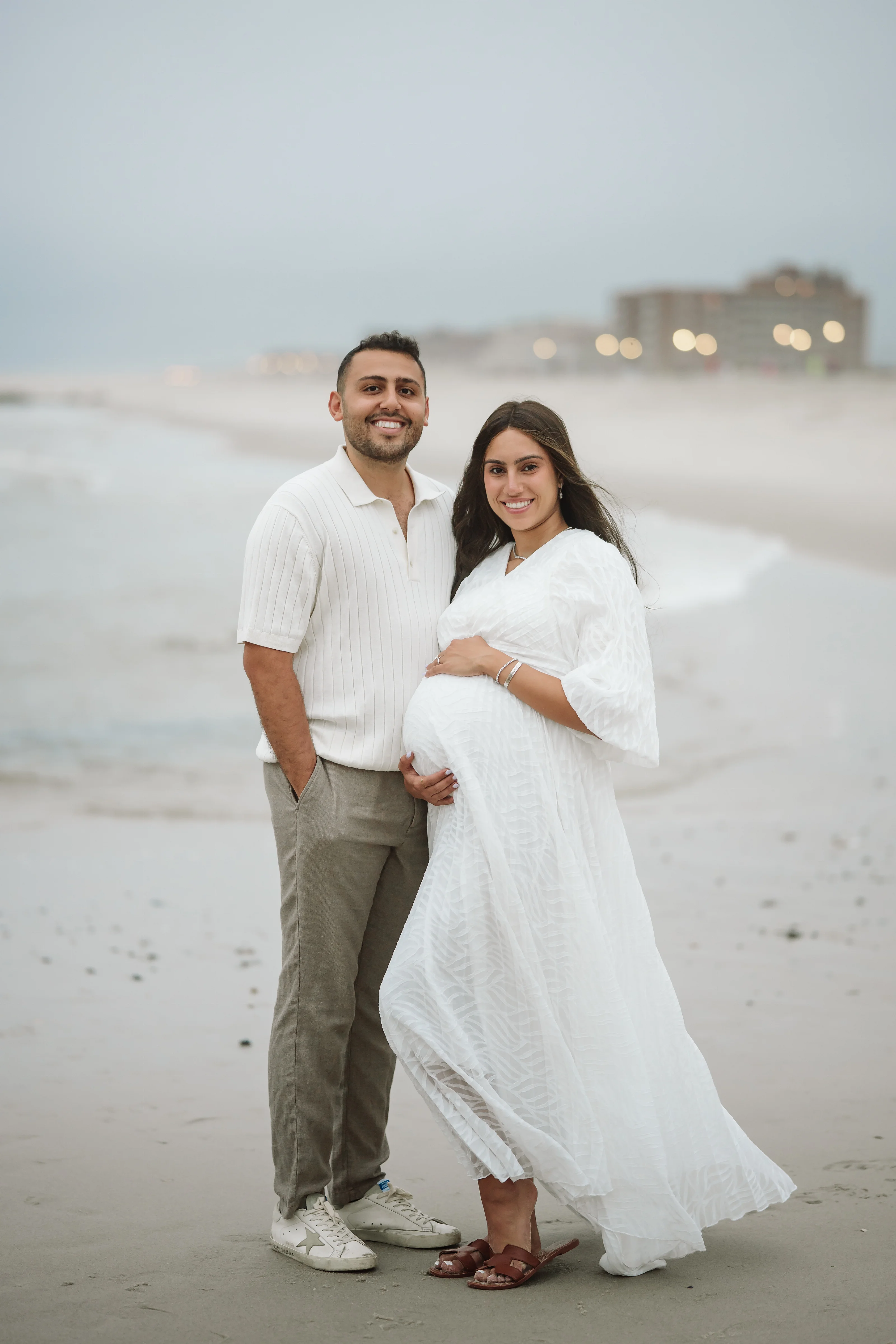 Couple posing on a sandy beach in white maternity attire, standing close with the father’s arm around the mother’s waist and both smiling at the camera.