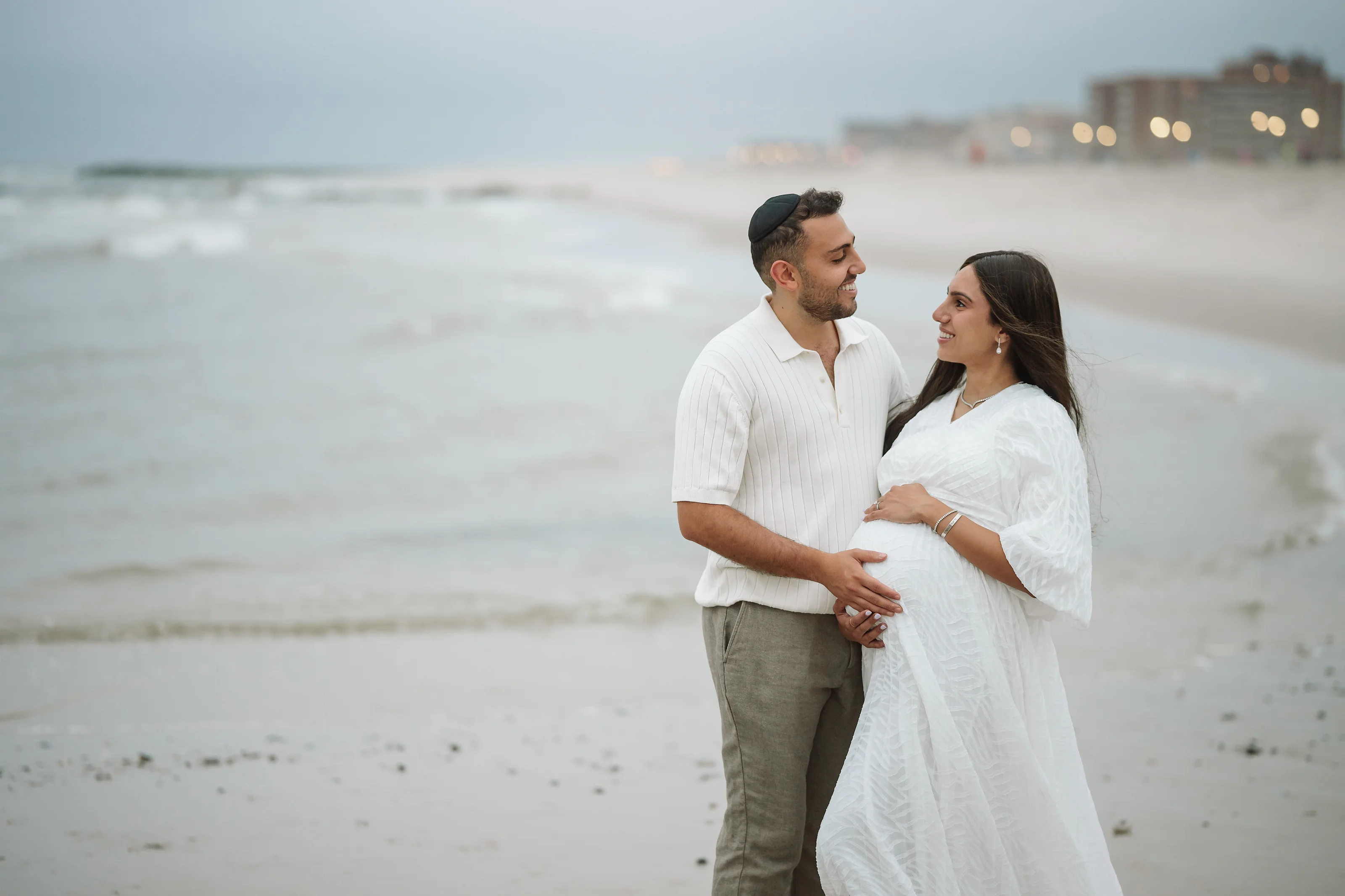 A man in a light button-up shirt and kippah holds his partner’s waist as she stands in a white flowing dress on a sandy beach with waves in the background.
