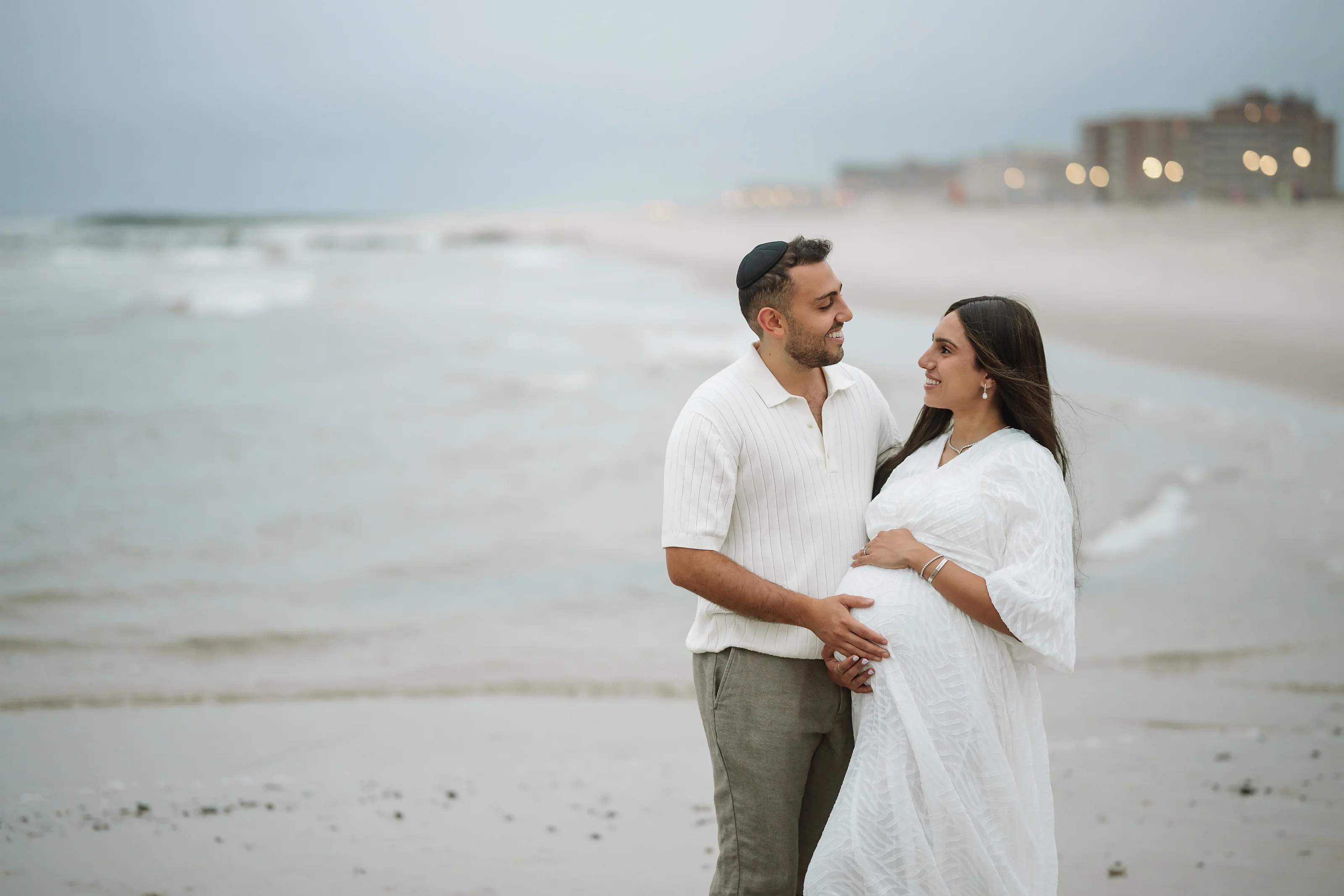 Maternity portrait of a couple standing on a sandy beach; the man in a white short-sleeve shirt and the woman in a flowing white dress, holding her belly while they smile at each other.
