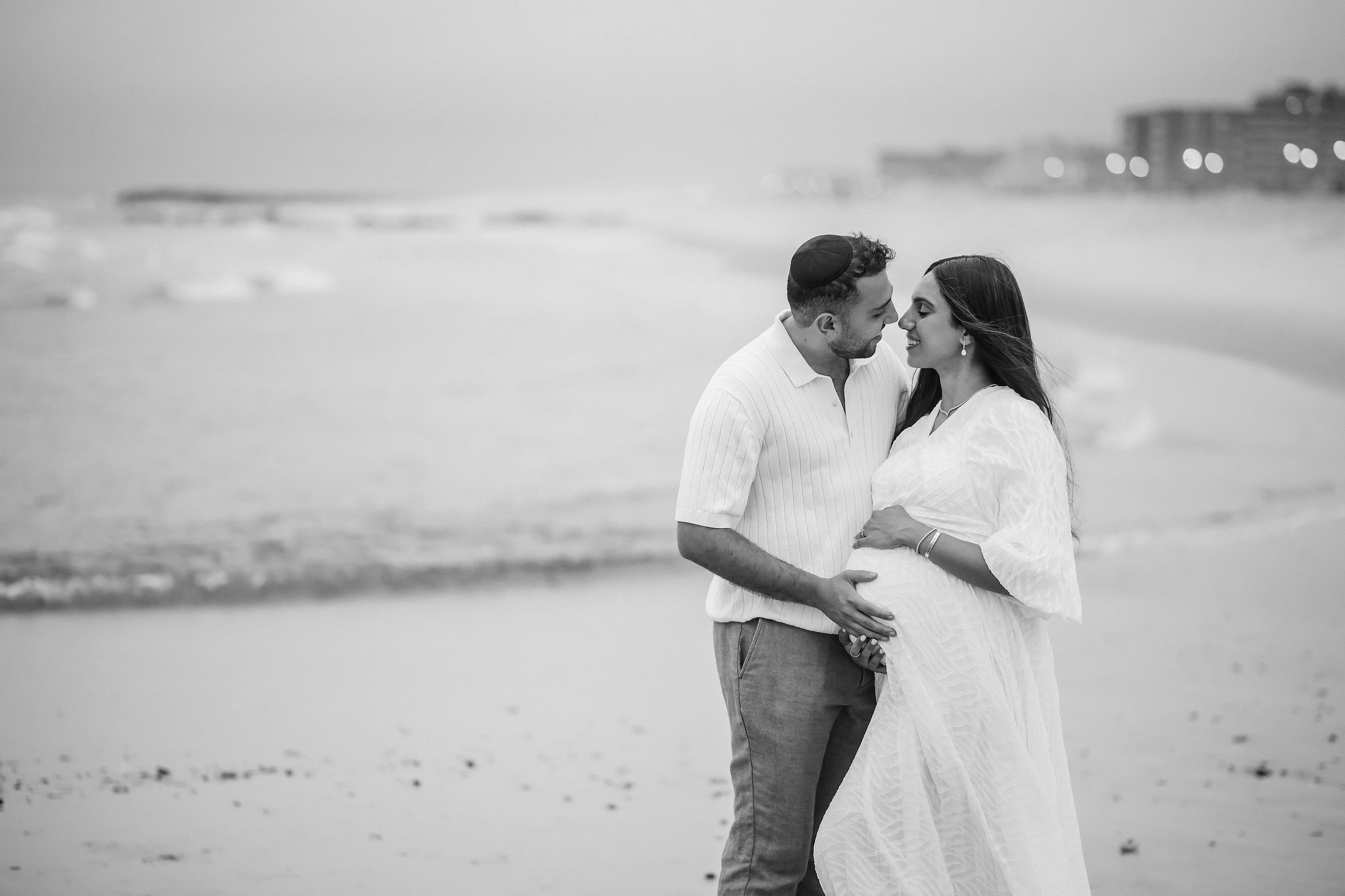 Black-and-white maternity portrait of a couple embracing by the water, with the woman in a white dress and the man in a light short-sleeve shirt, their faces turned toward each other.