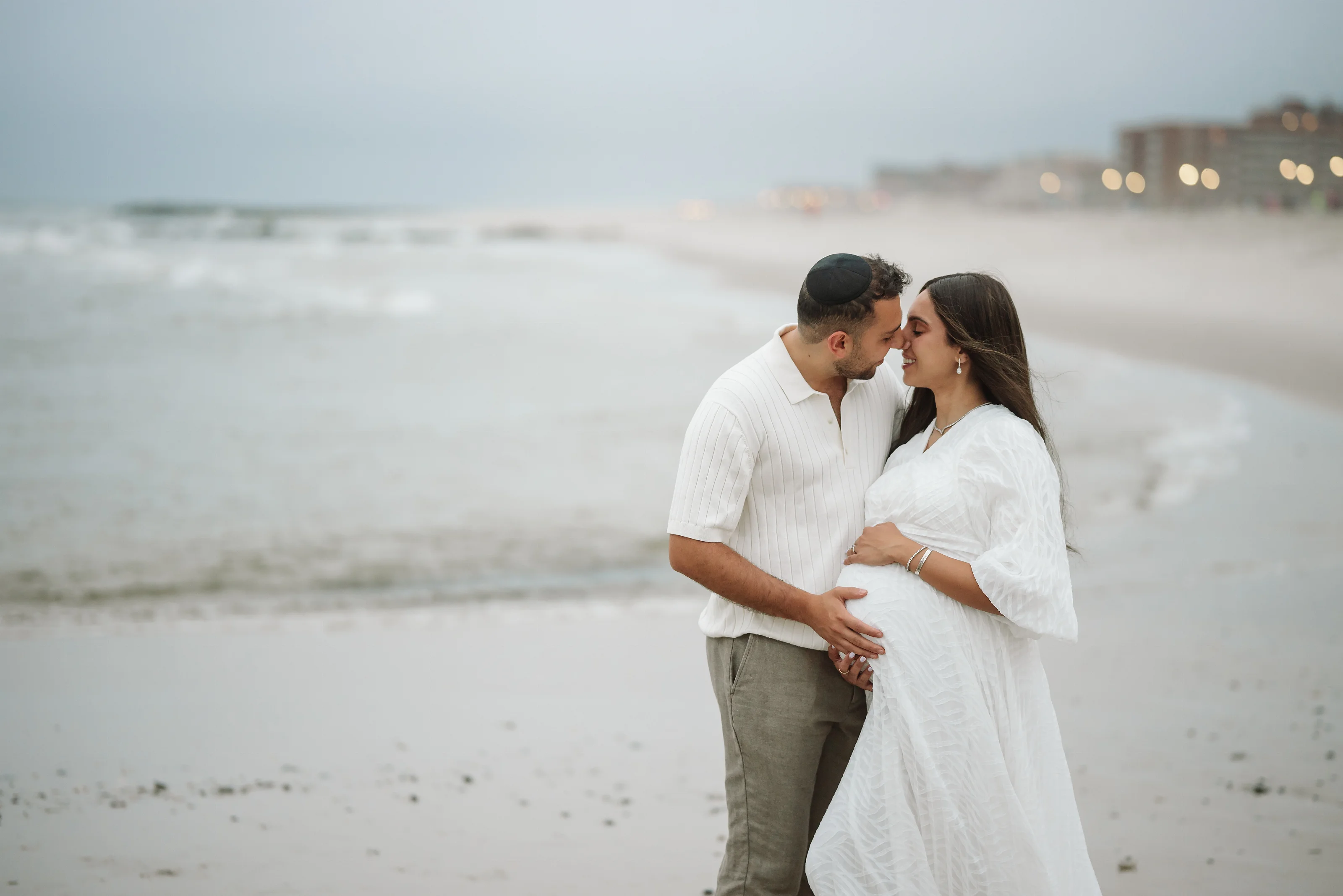 A couple in white and light neutral clothing stands on a beach, embracing and kissing while one partner cradles an expectant belly.