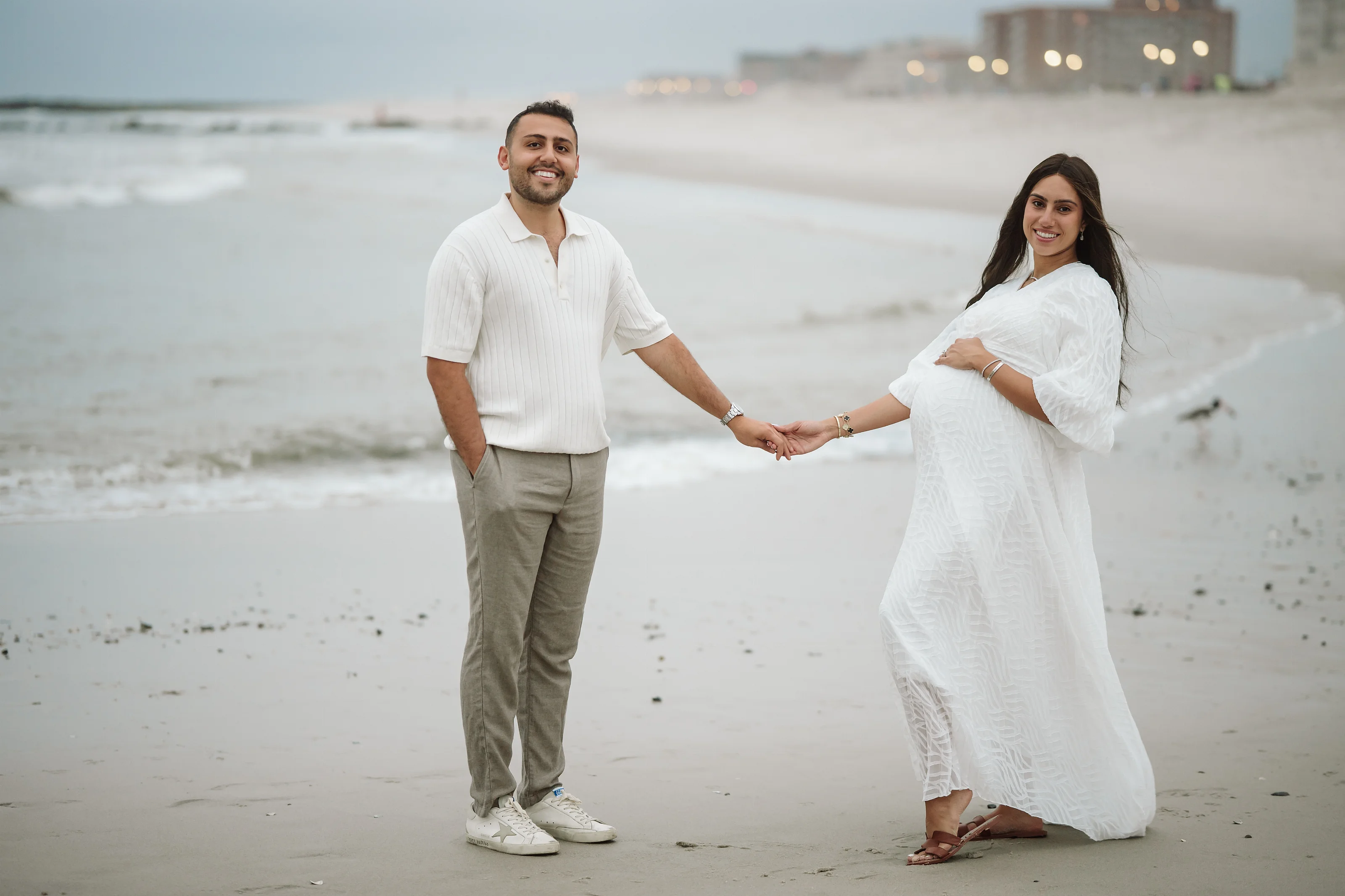 A man in a white short-sleeve shirt and light pants and a woman in a flowing white maternity dress stand on a beach holding hands, smiling toward the camera with the ocean behind them.