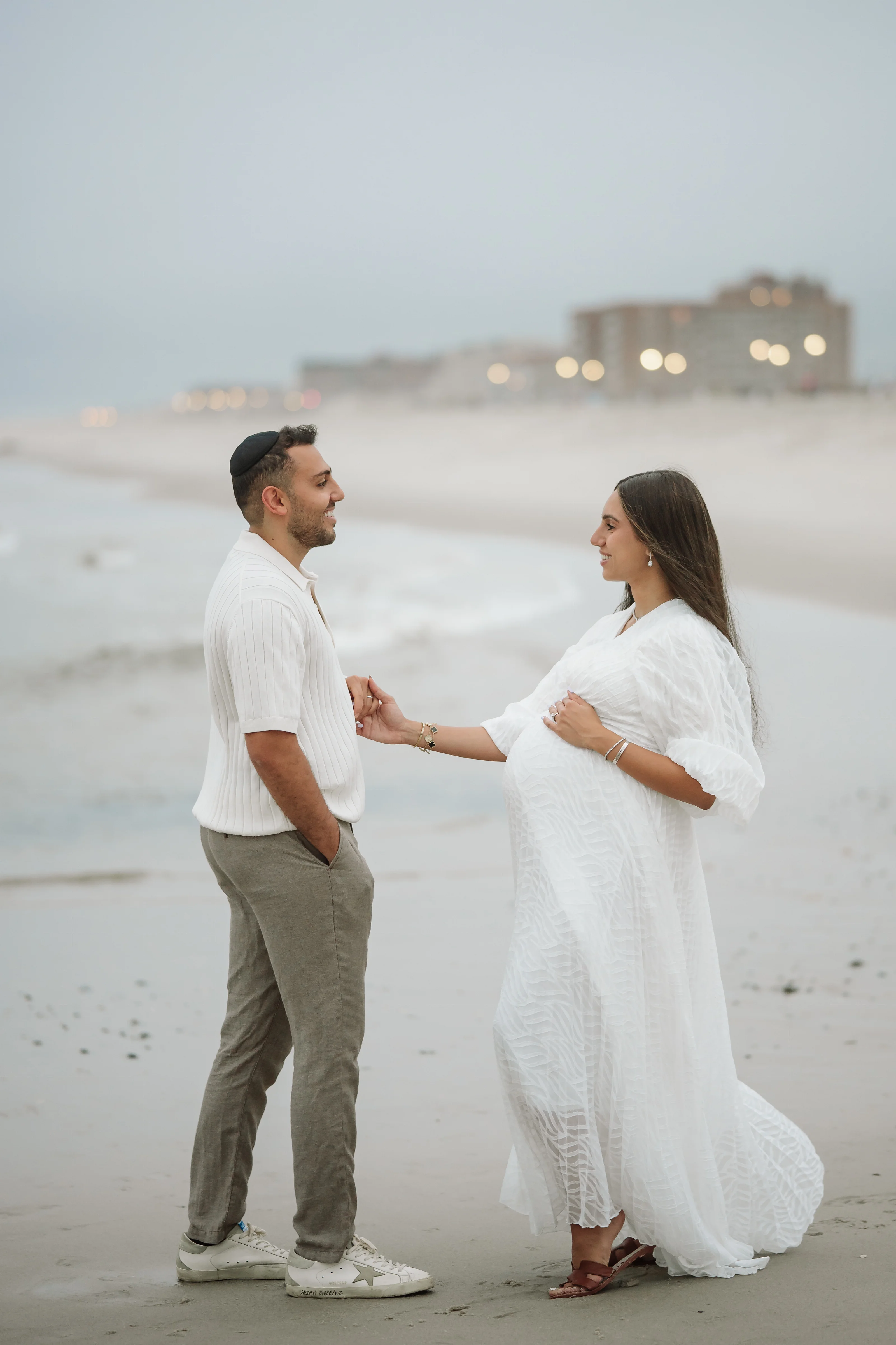 A couple in white and light outfits stands on a beach holding hands, facing each other and smiling as the sky fades to dusk.