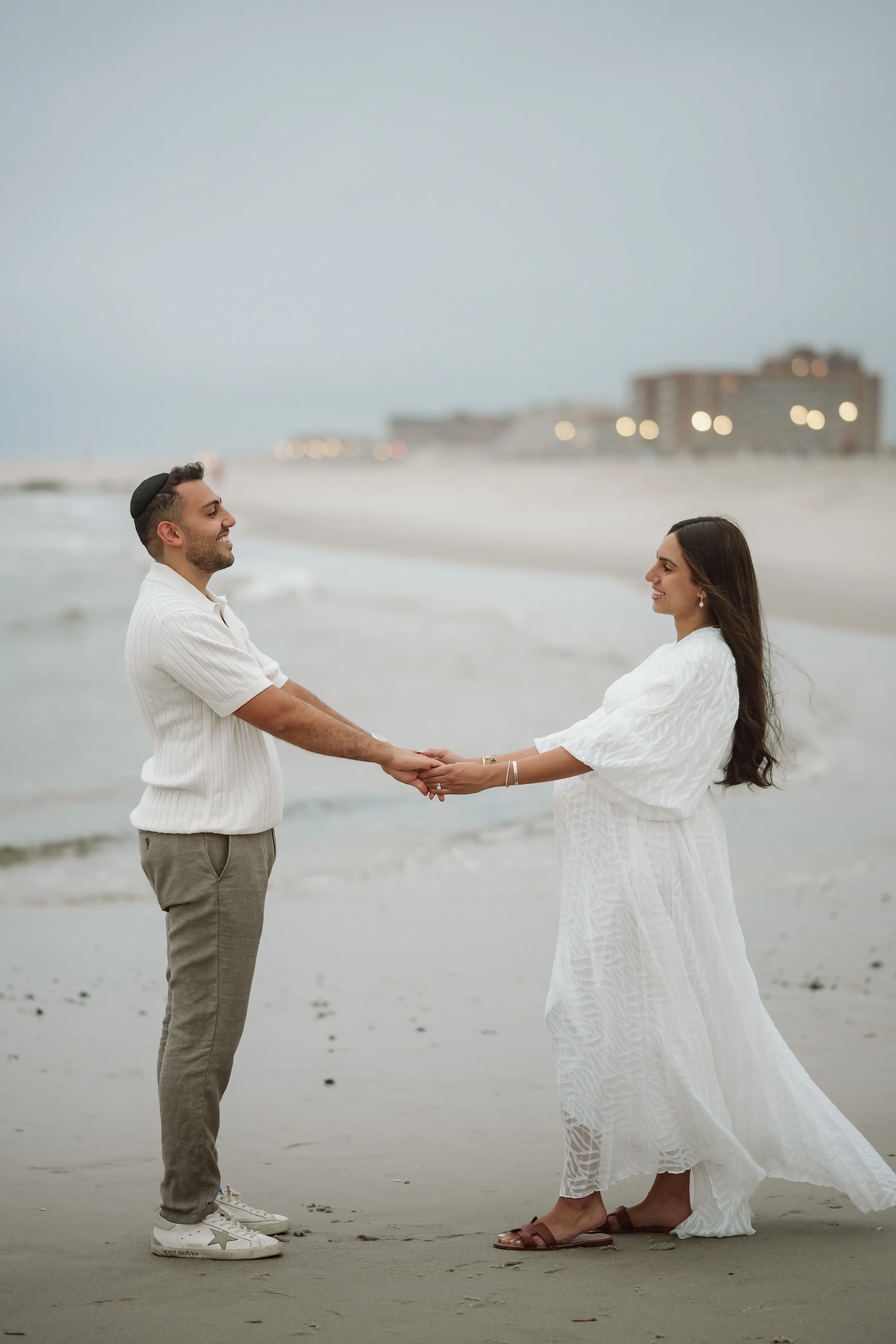 Expecting couple holding hands on a quiet beach at dusk, with the woman in a flowing white dress and the man in a light shirt and olive pants.