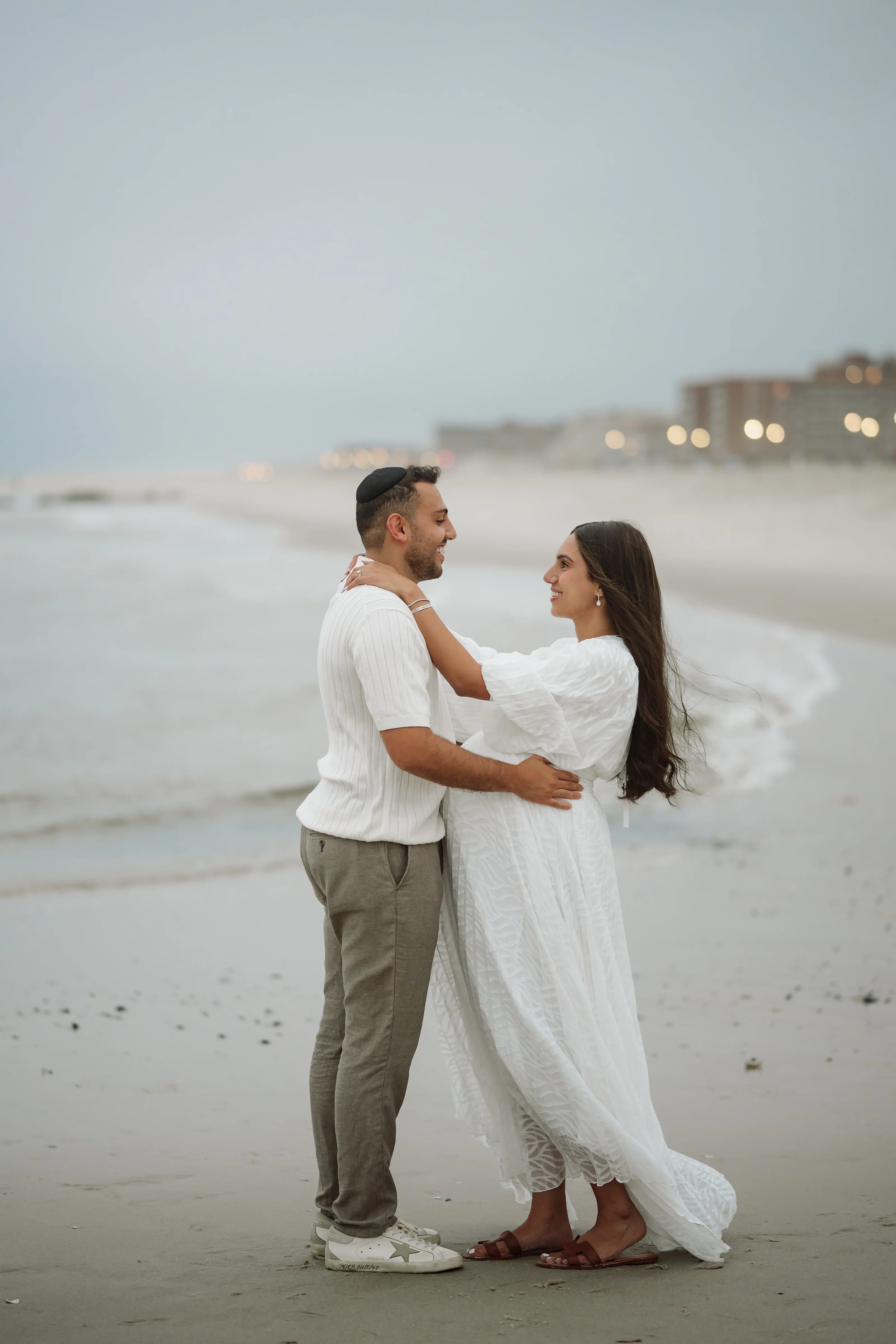 A couple stands close in a beach embrace at golden hour, with the woman in a flowing white dress and the man in a white shirt and neutral pants.