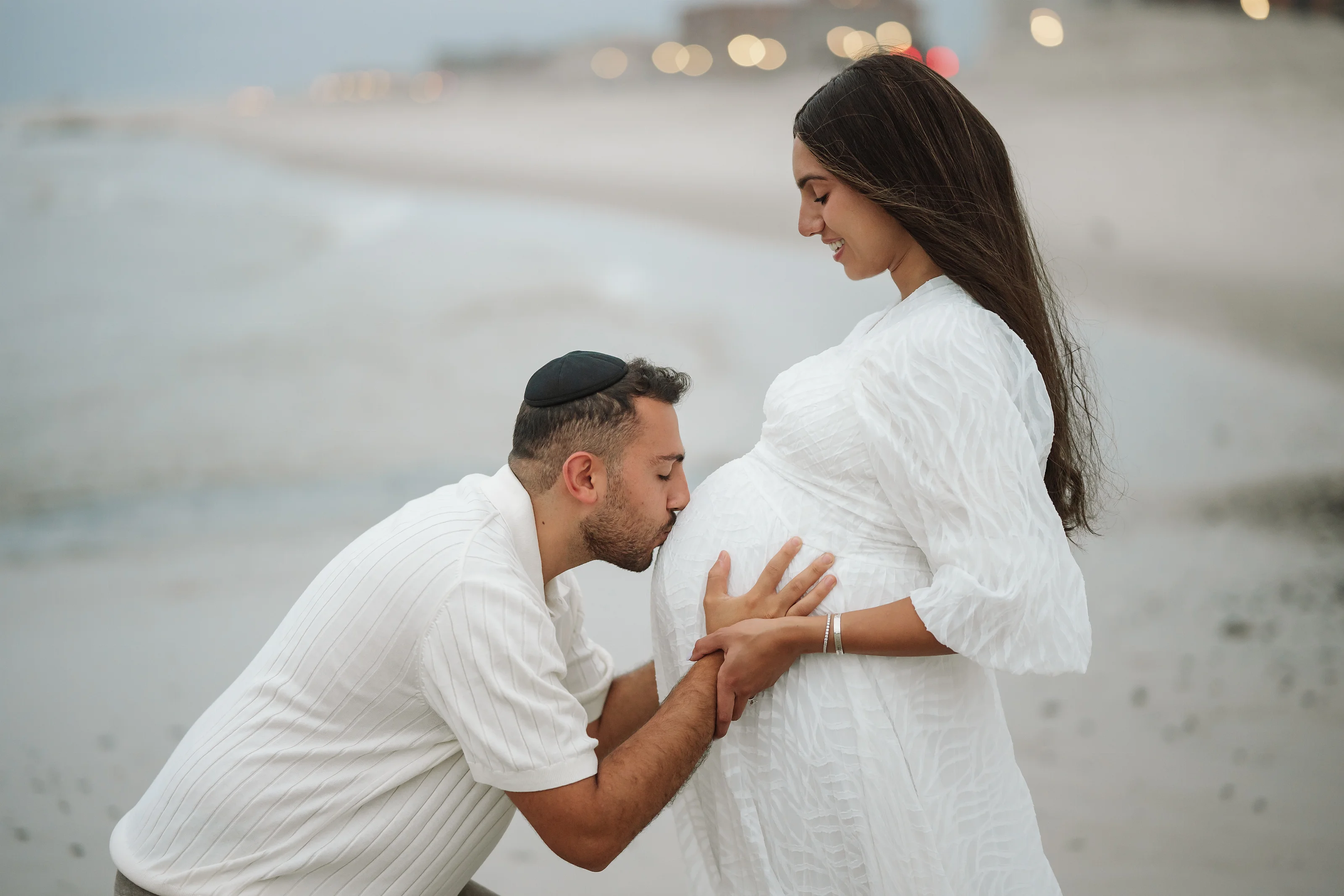 A woman in a flowing white dress stands by a kneeling partner in a white shirt on a beach, their hands held together as they look down.