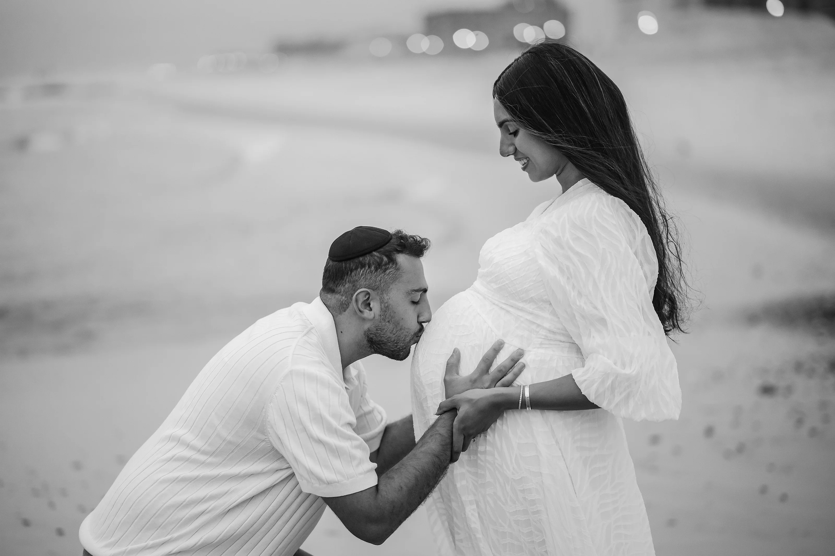 Black-and-white maternity portrait of a pregnant woman in a light textured dress standing in profile, with her partner kneeling and gently holding her belly against a softly blurred background with bokeh lights.