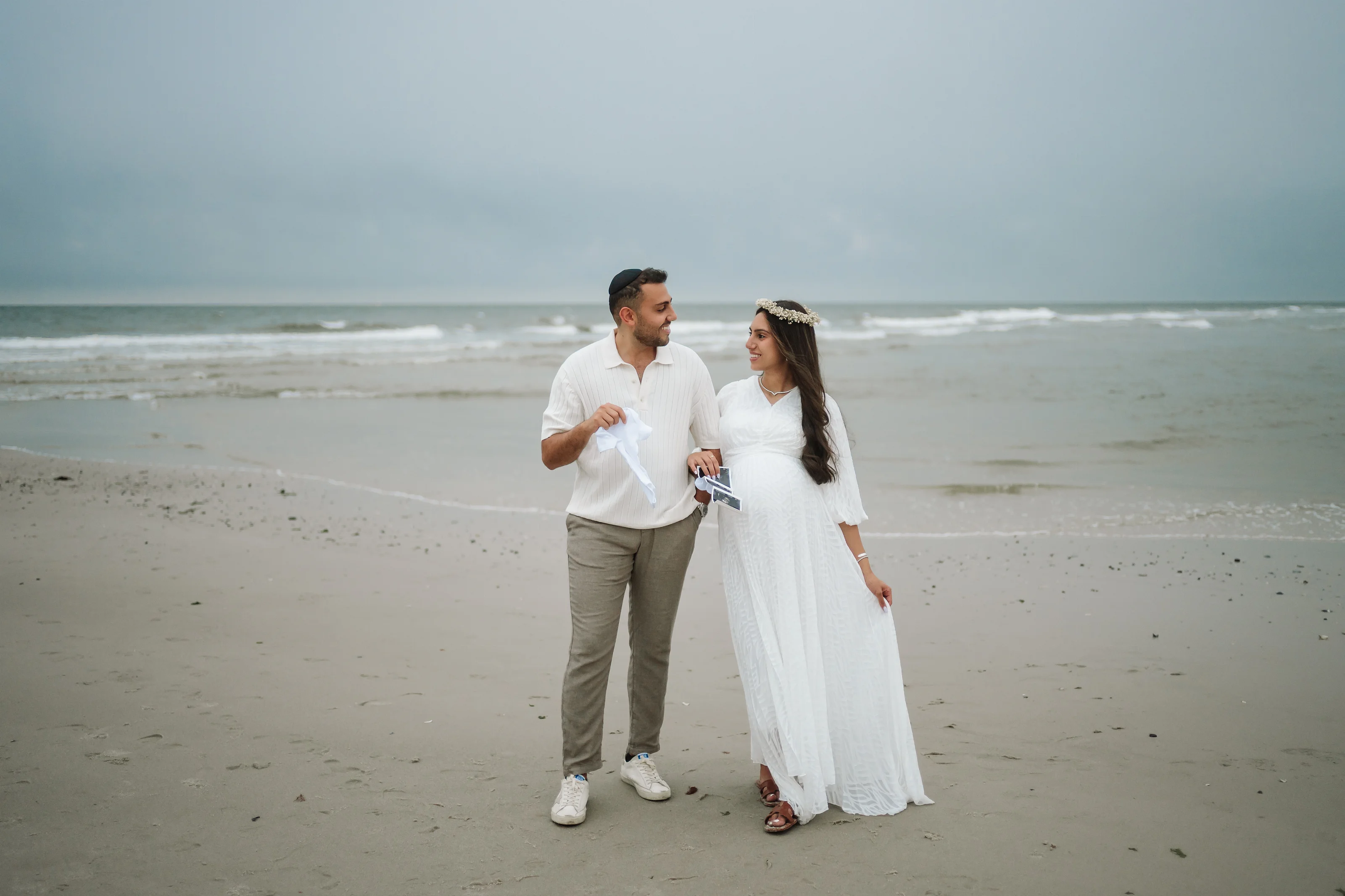 A couple stands on a sandy beach near the shoreline, the woman in a flowing white dress with a headpiece and the man in a white shirt and light trousers, facing each other with relaxed smiles.