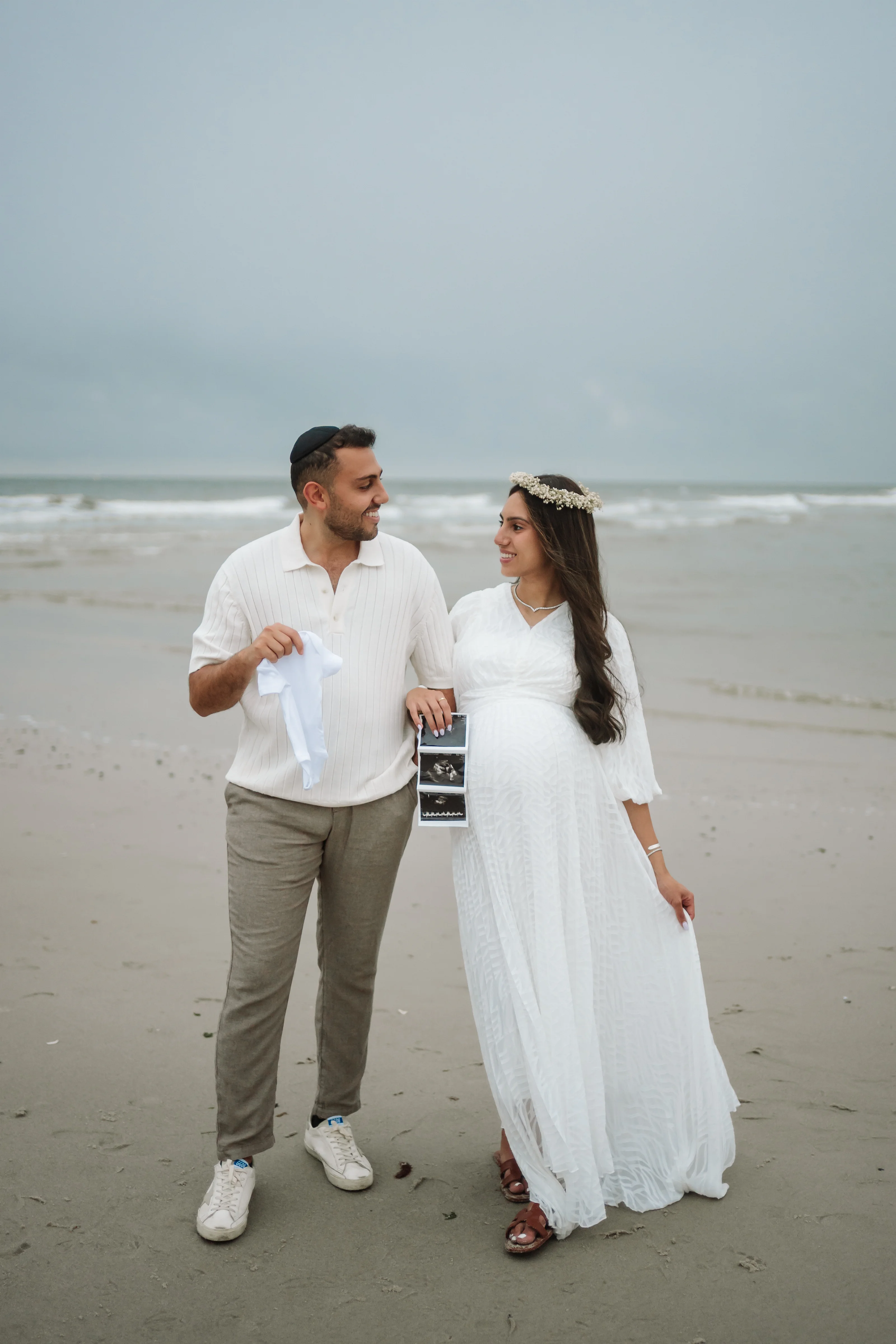 Couple in white outfits standing on a sandy beach near the ocean, with the bride wearing a white dress and floral headpiece and the groom in a white shirt and gray pants, smiling at each other in soft overcast light.