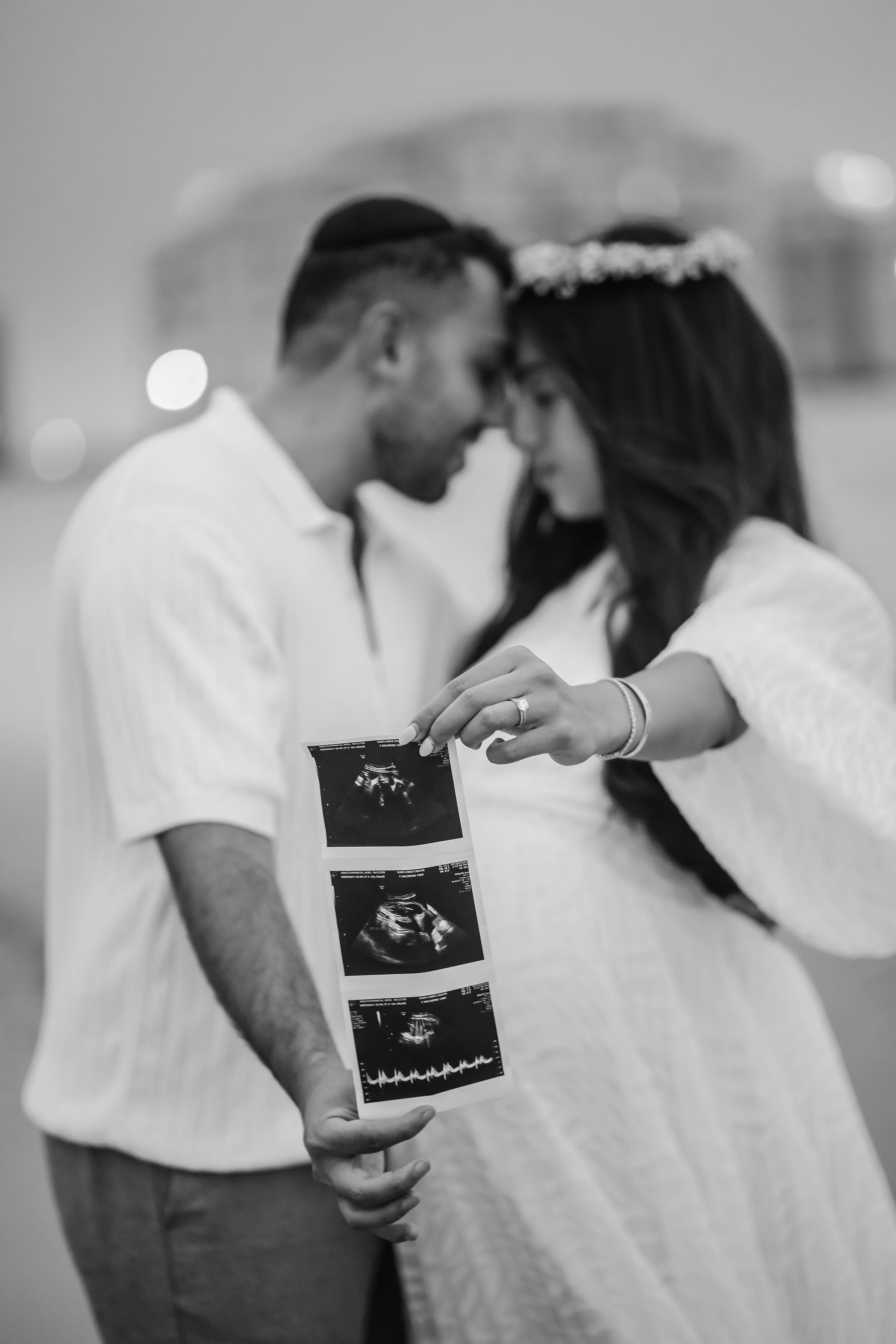 Black-and-white maternity portrait of a couple kissing while one person holds ultrasound printouts in the foreground, with the other’s hands resting at their shoulder in a softly lit interior.