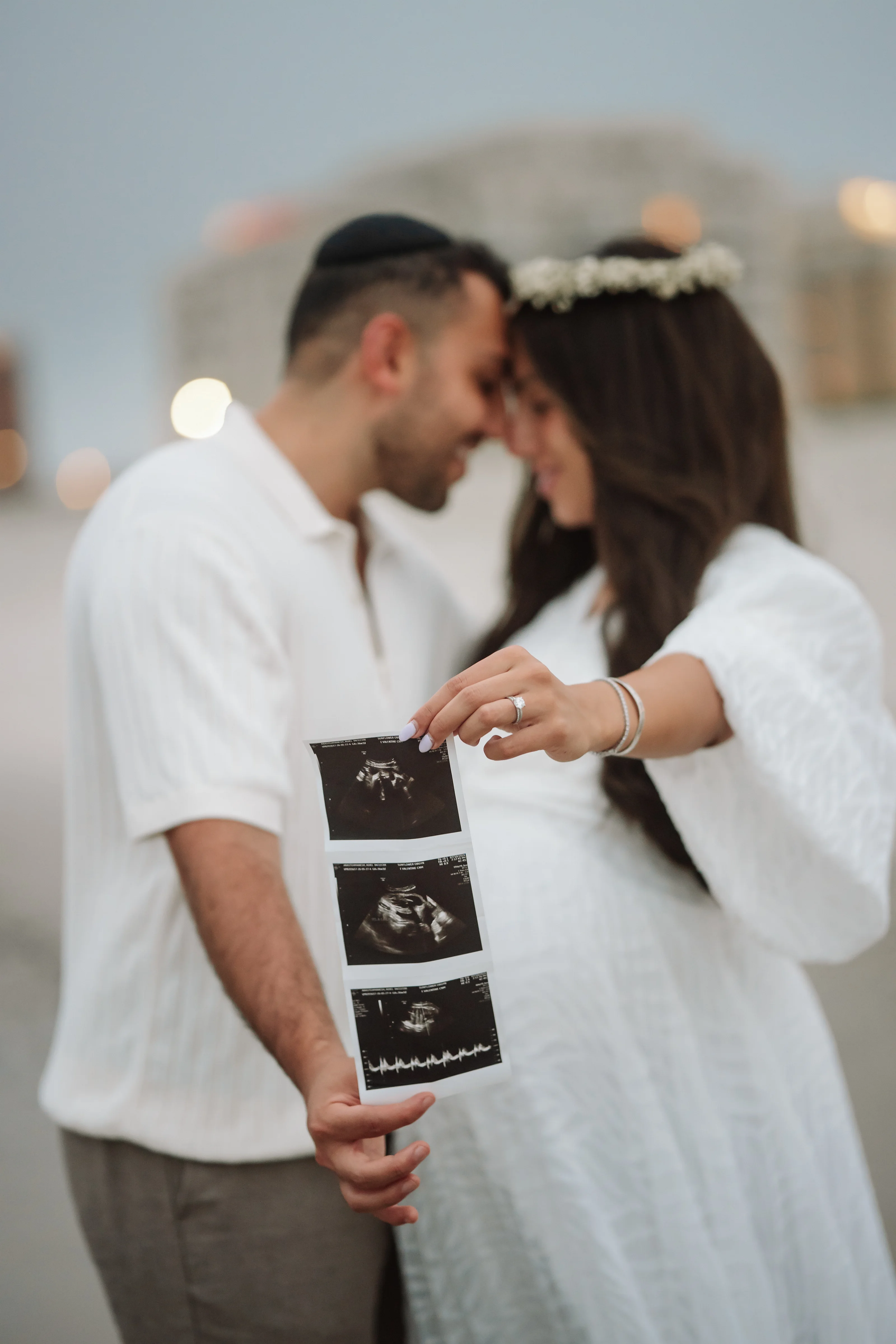 A couple in white stands close and forehead-to-forehead while holding a printed ultrasound photo set; the woman wears a light floral headband as they smile softly in a softly lit indoor setting.