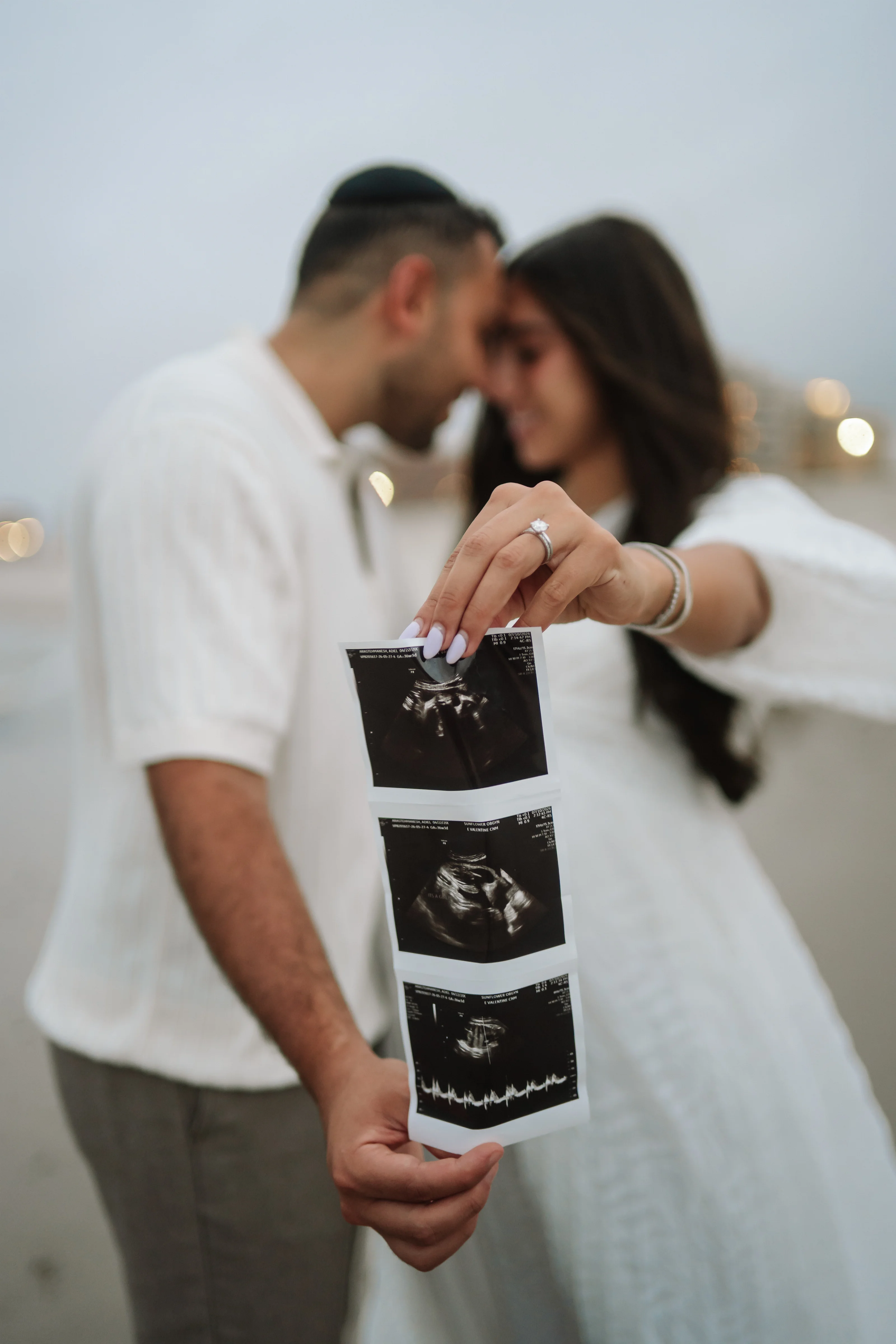 Couple in light clothing sharing a close kiss as ultrasound printouts are held up in the foreground against a soft, softly lit background.