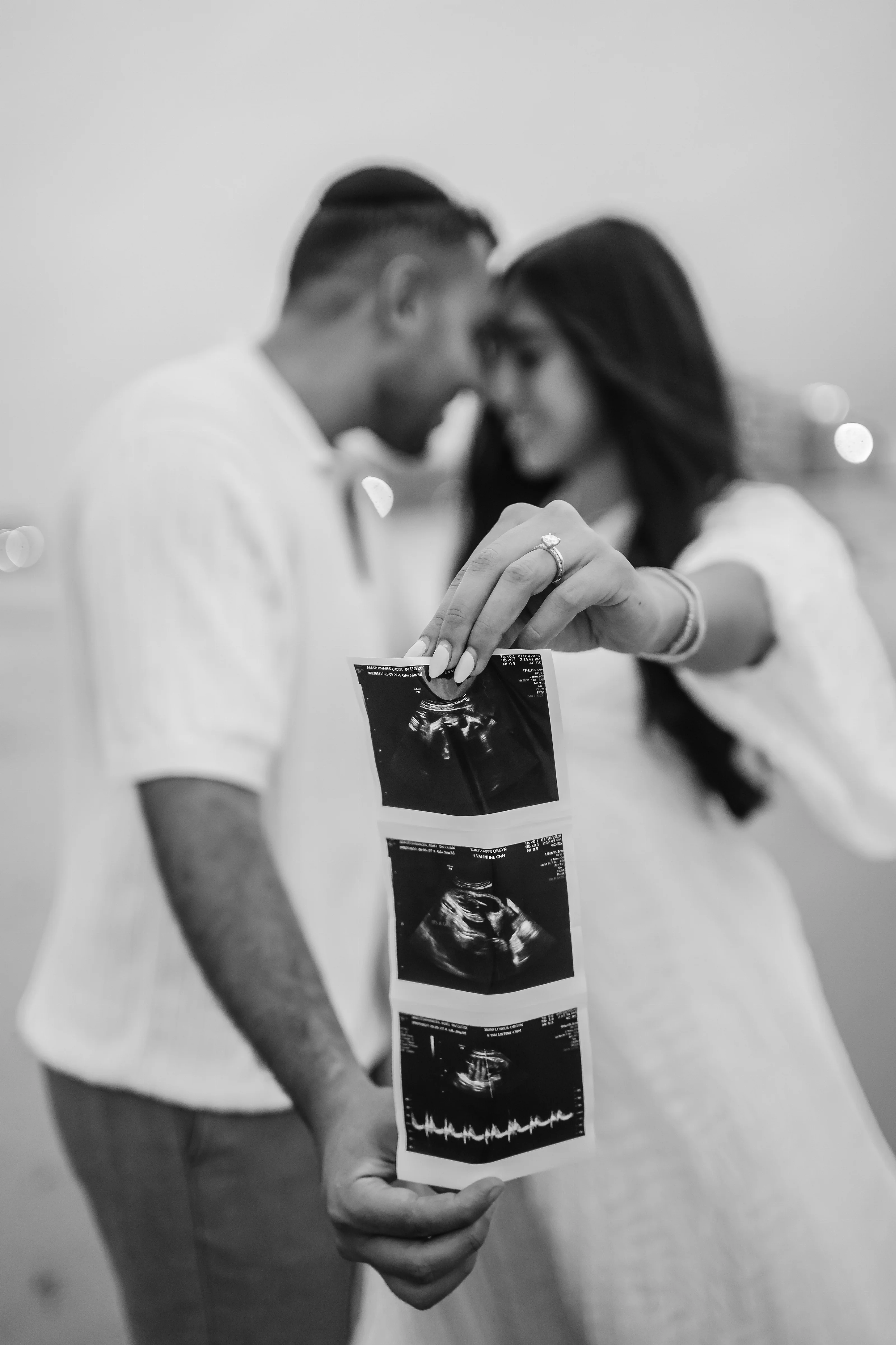 Black-and-white maternity portrait of a couple leaning close, with ultrasound printouts held in the foreground and a wedding band visible on the mother’s hand.