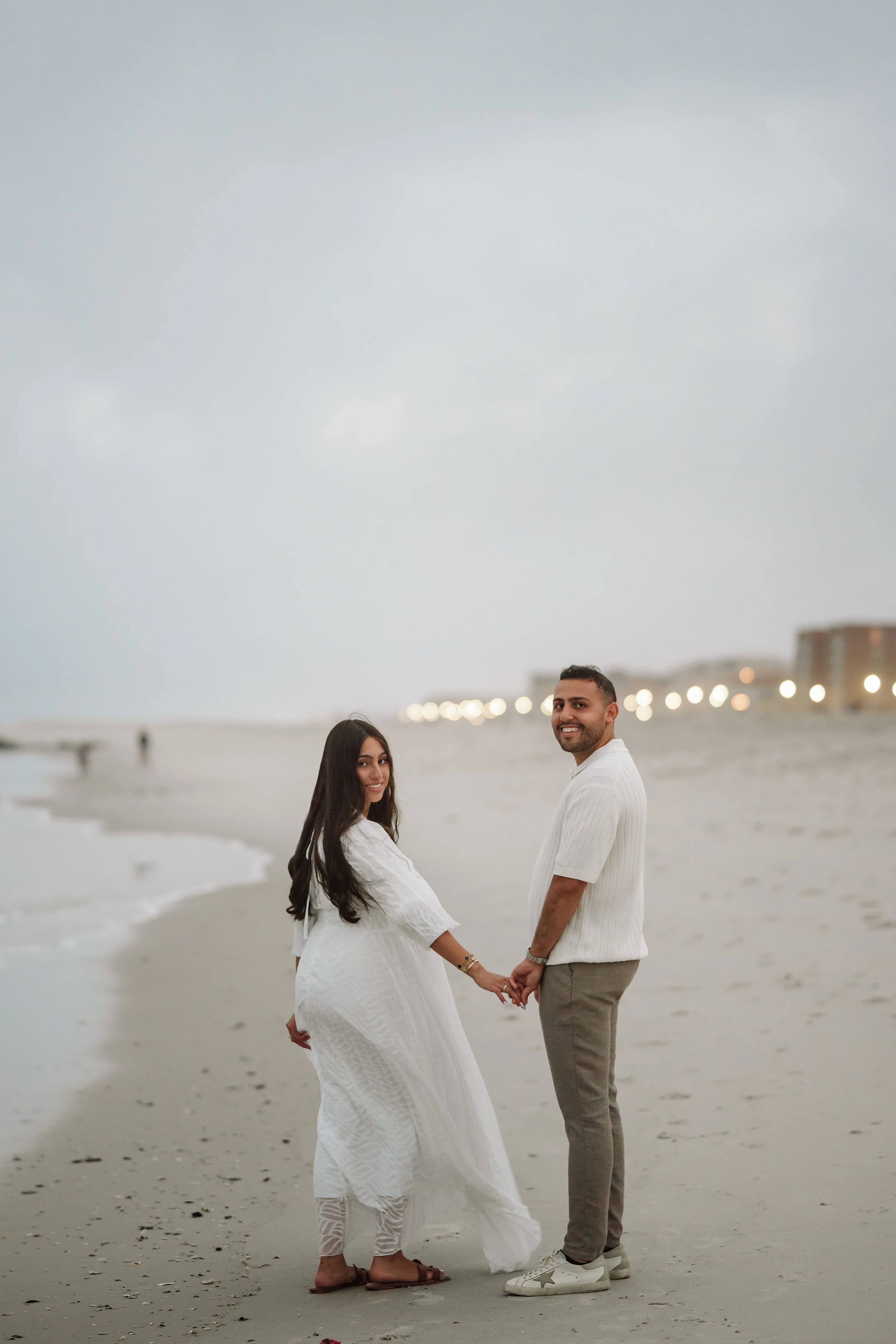 A couple in flowing white outfits holds hands and walks on a sandy beach at dusk with soft lights glowing in the distance.