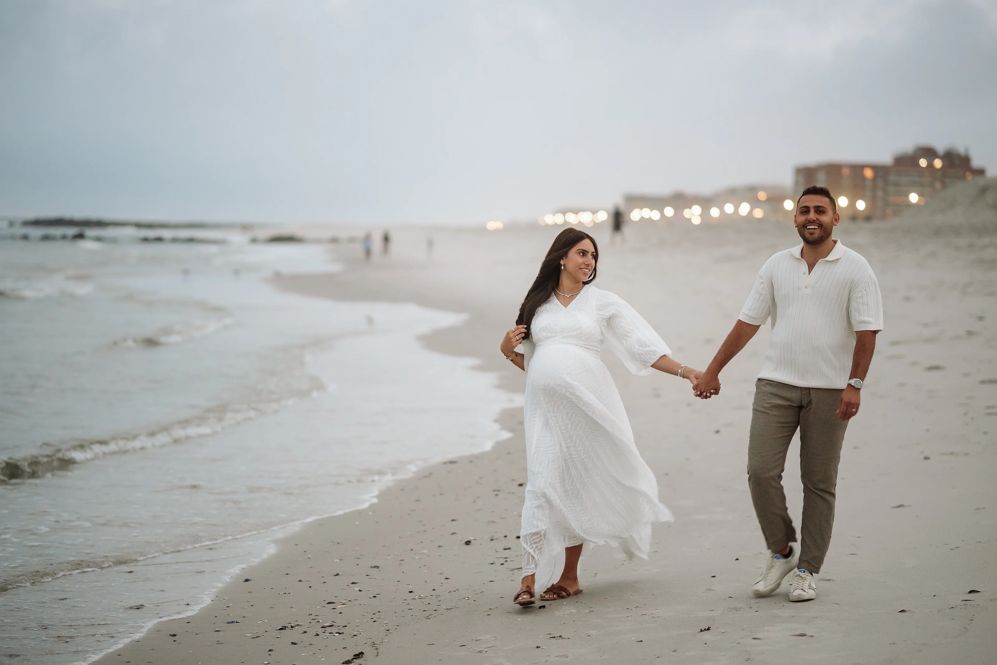 A couple holding hands on a sandy beach at dusk, the woman wearing a flowing white dress and veil and the man wearing a light knit shirt and casual pants.