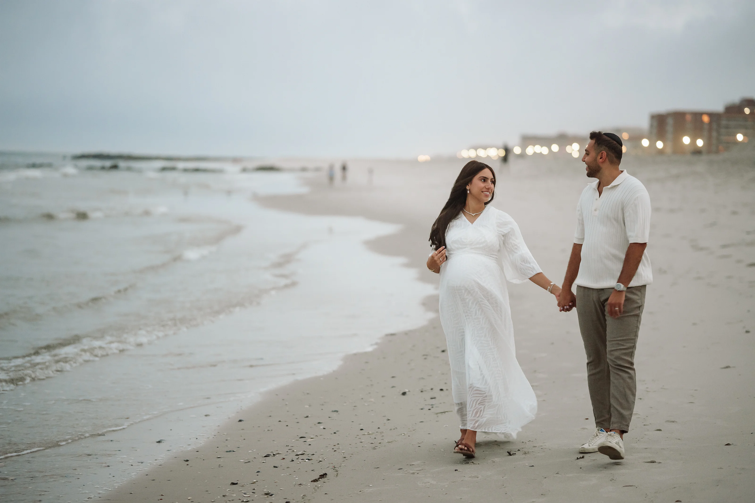 A woman in a white flowing maternity dress and a man in a white shirt and tan pants walk hand-in-hand on a sandy beach near the shoreline at dusk.