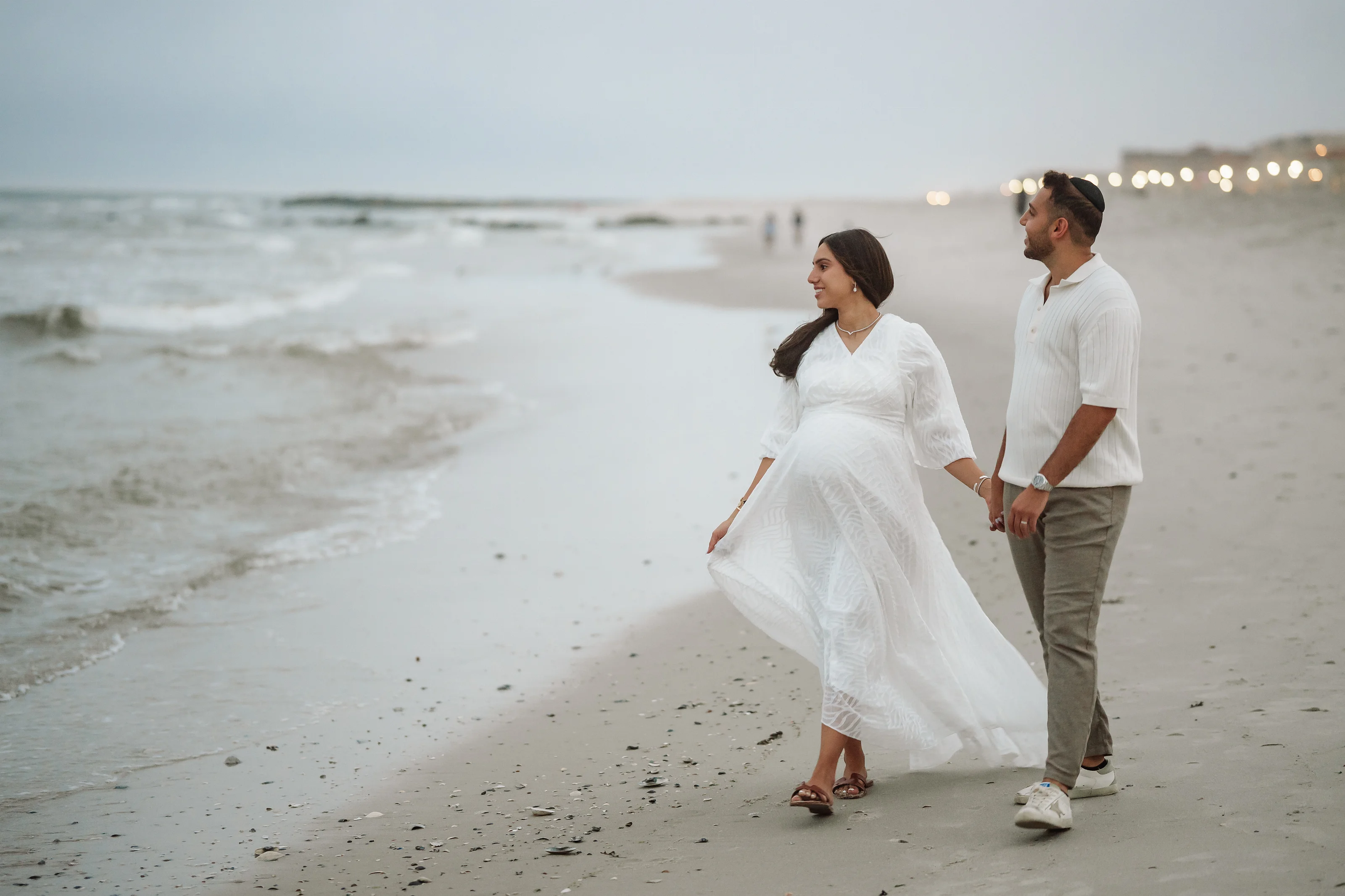 Couple on a sandy beach at dusk walking hand-in-hand, bride in a flowing white maternity dress and groom in a light button-down and gray trousers.