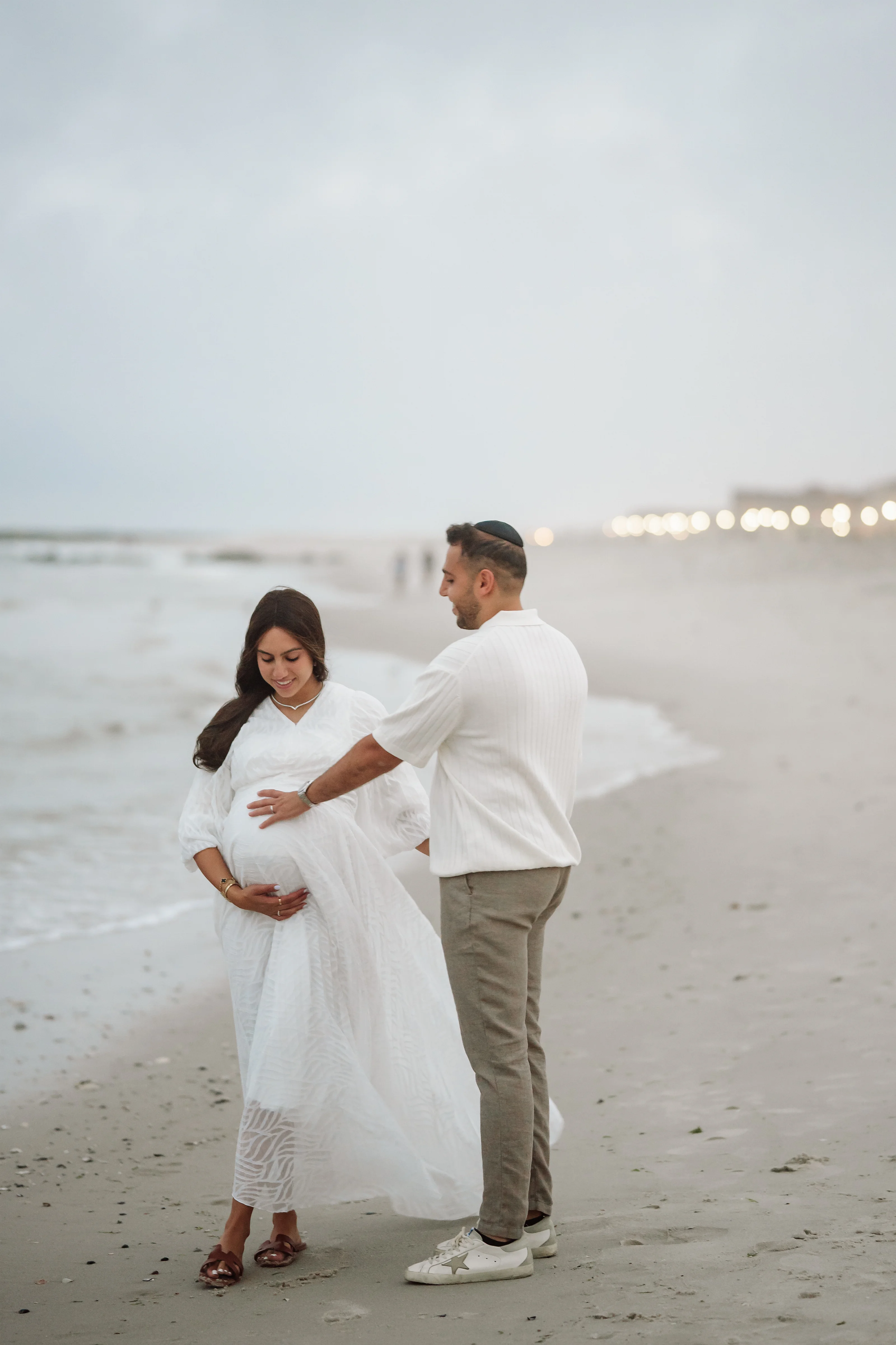 Expectant couple standing on a sandy beach at dusk, wearing light white outfits, with hands resting over the baby bump while the ocean and distant string lights glow behind them.