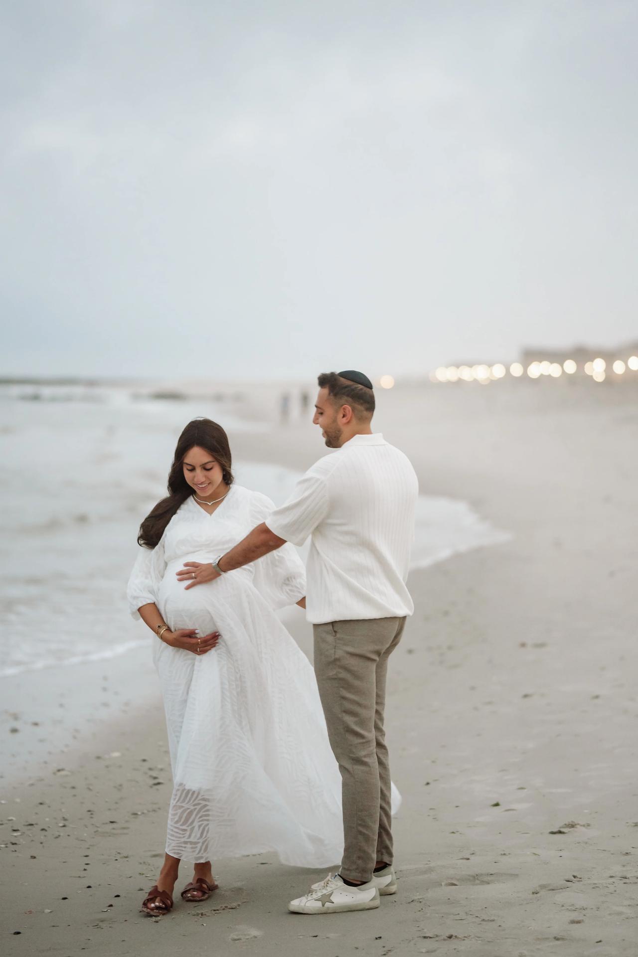 Expectant couple standing on a sandy beach at dusk, wearing light white outfits, with hands resting over the baby bump while the ocean and distant string lights glow behind them.