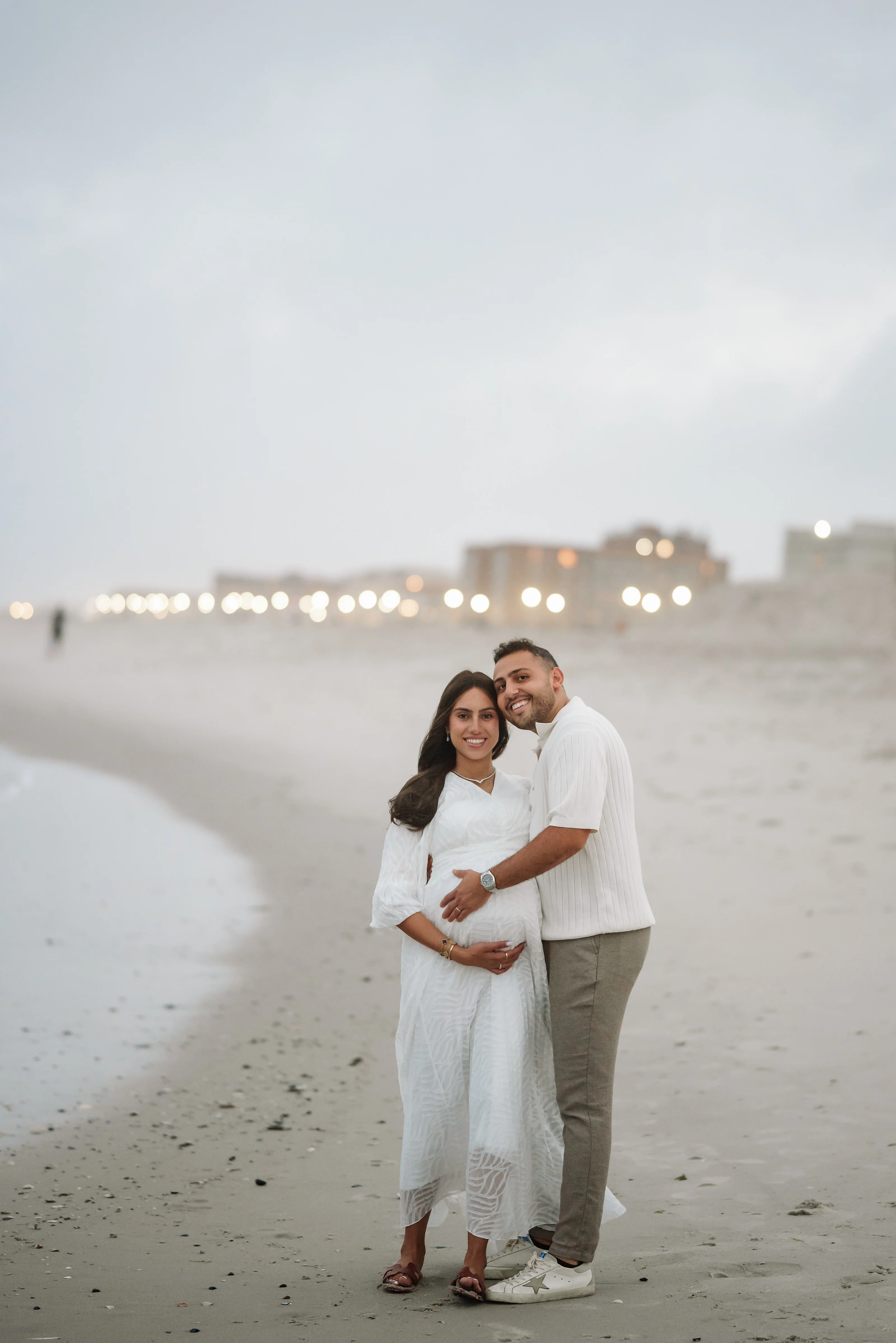 Outdoor maternity portrait of a couple standing close together on a beach at dusk, with the expectant mother in a white dress and the expectant father in a white shirt and beige pants, warm lights glowing in the distance.
