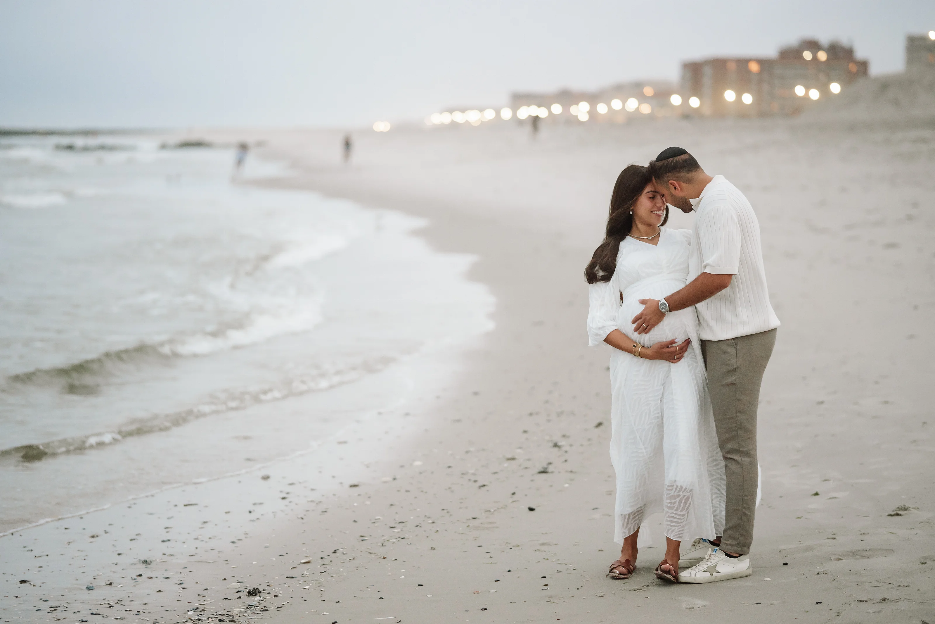 Couple in matching white maternity outfits standing on a sandy beach at sunset, embracing and smiling with the ocean and distant lights in the background.