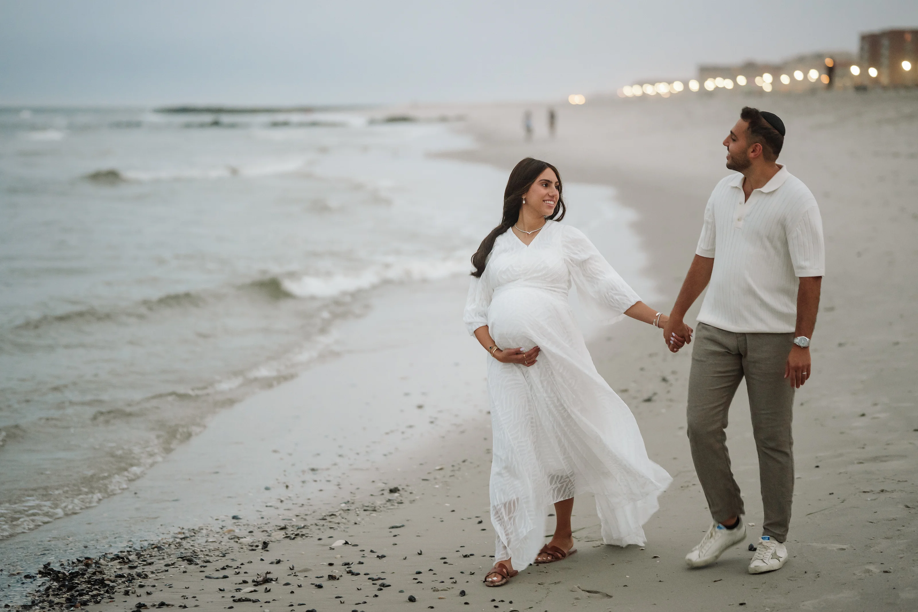 Woman in a flowing white maternity dress and necklace and a man in a white shirt and gray pants walking hand-in-hand along a sandy beach at dusk, with ocean waves behind them.