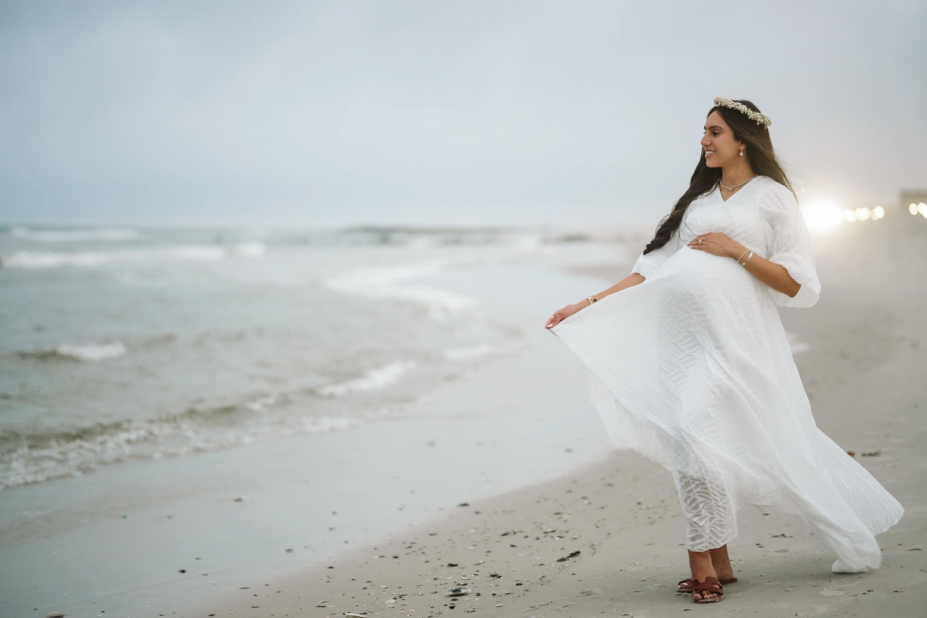 A pregnant woman in a flowing white maternity dress and headpiece stands barefoot on a beach with her hand resting on her belly, gazing to the side near the shoreline.