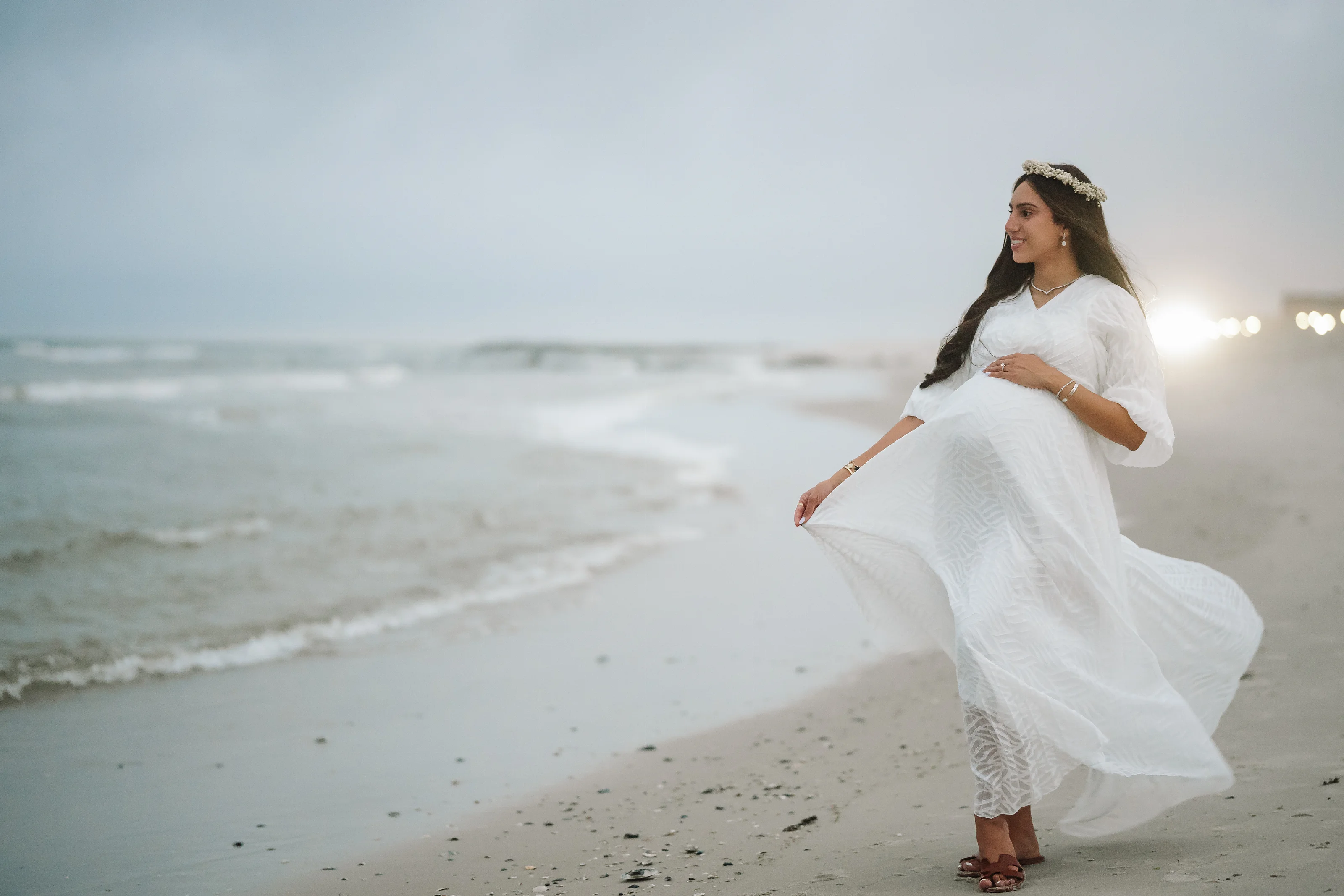 Woman in a flowing white maternity dress and sandals walking along a beach shoreline at sunset, one hand resting on her belly and looking to the side.