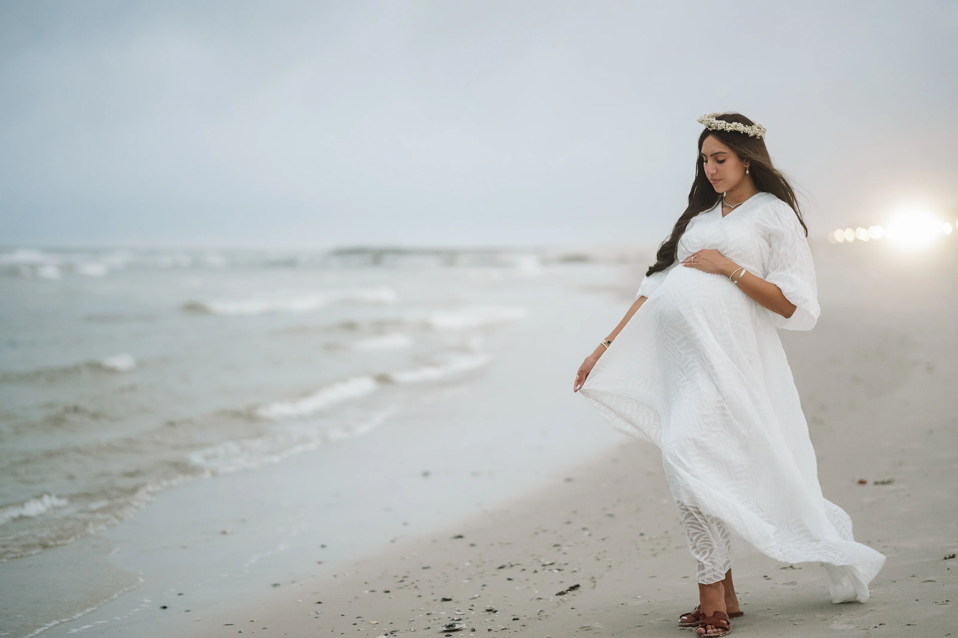 A woman in a white maternity dress and matching headpiece walks on a sandy beach near the ocean, holding her belly at sunset light.