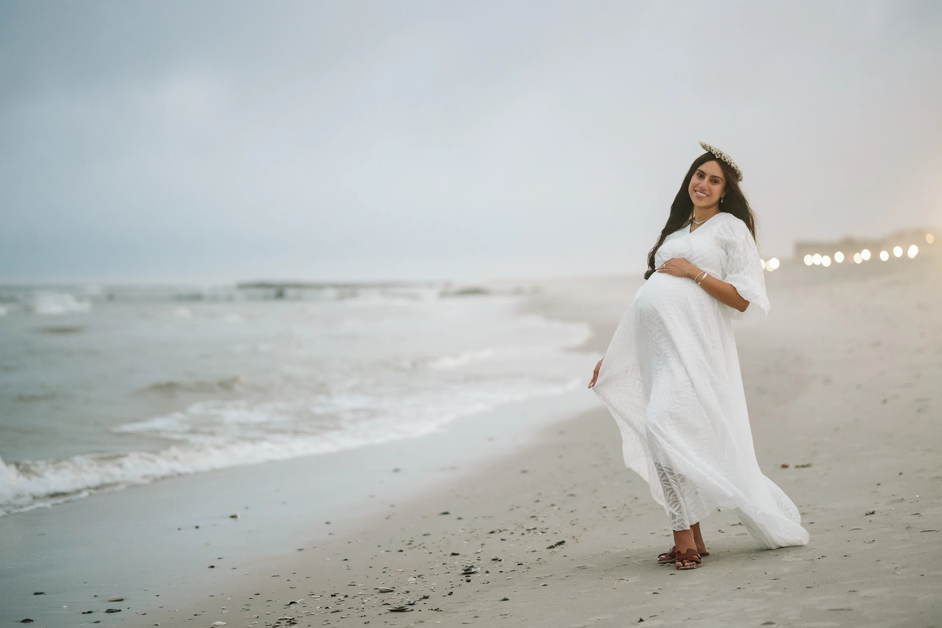 Woman in a flowing white maternity dress standing on a beach near the shoreline, one hand resting on her belly, with string lights in the distance.