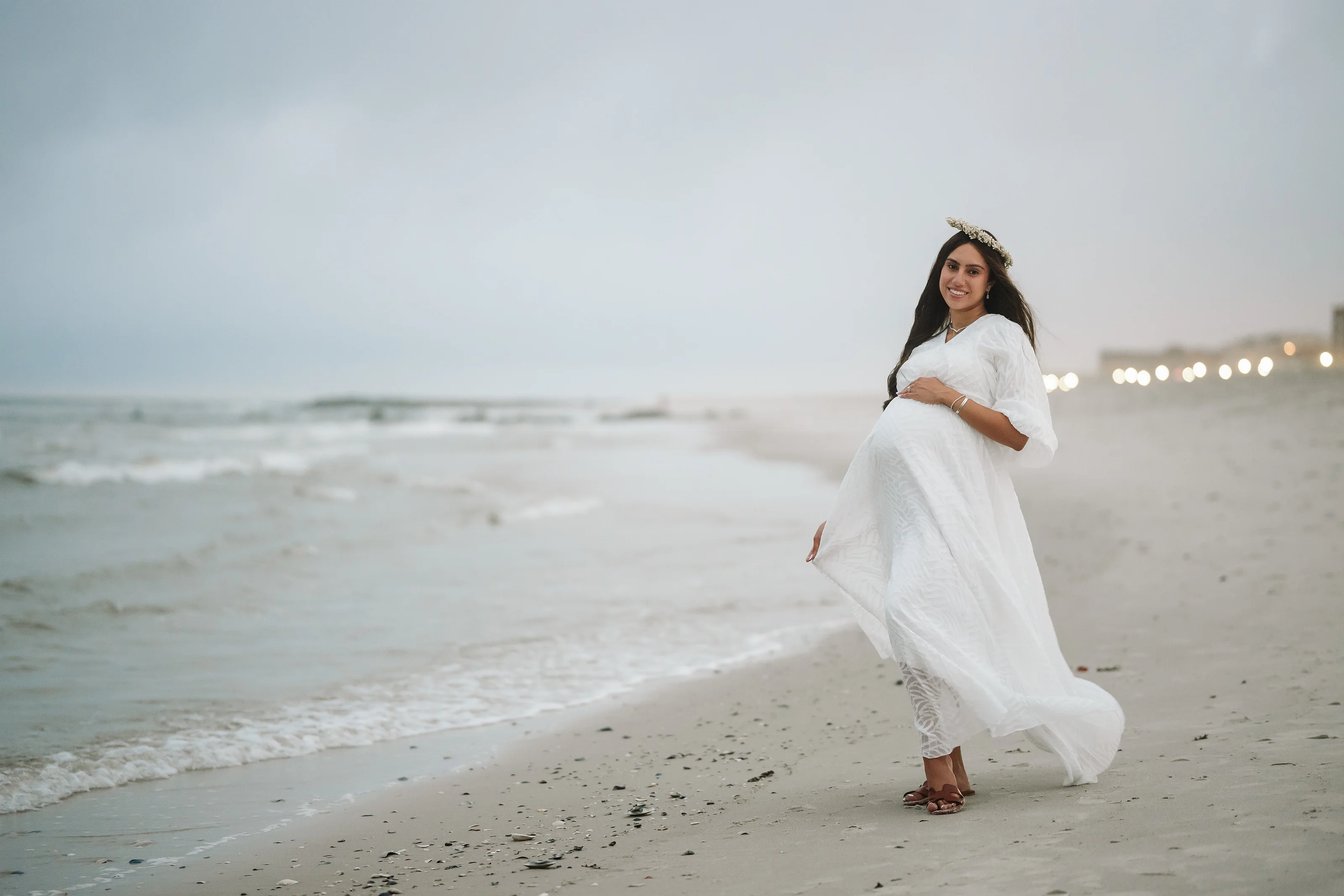 Woman in a flowing white lace maternity dress and floral headpiece posing on a sandy beach at dusk with soft string lights in the distance, smiling while standing and turning slightly.
