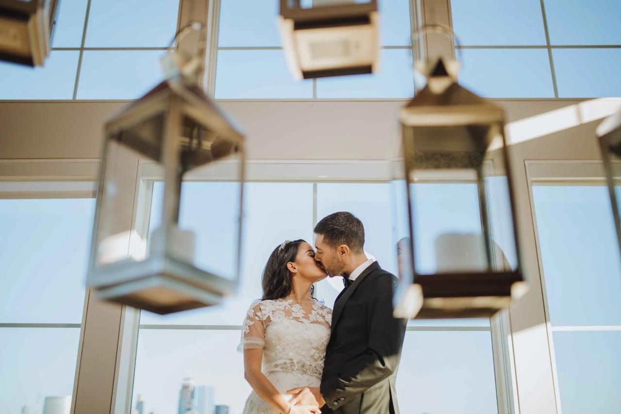 A bride and groom kiss while facing each other in close embrace against tall glass windows inside a bright venue.