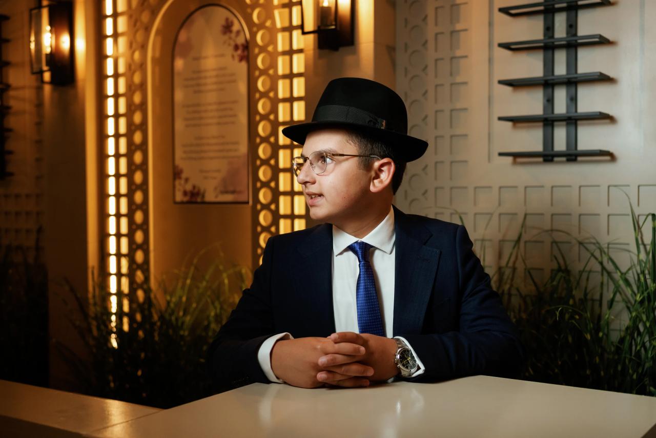 Portrait of the Bar Mitzvah boy in a navy suit, black hat and glasses at Hilton Garden Inn Springfield, NJ