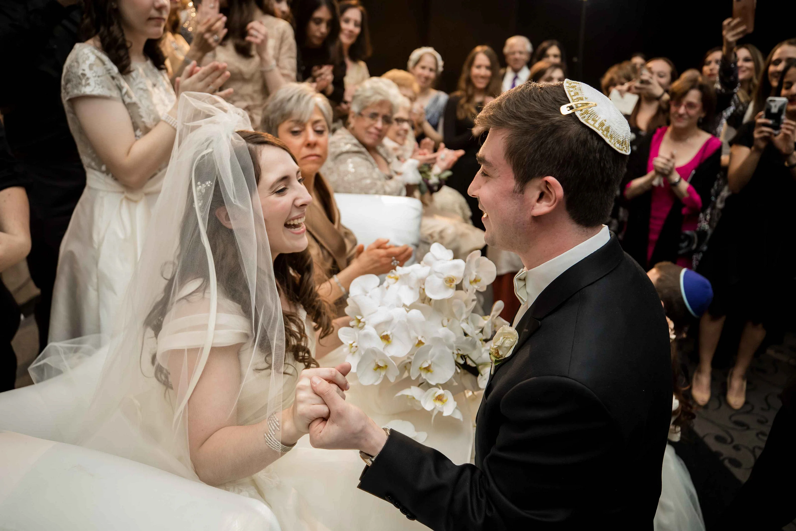 Bride and groom facing each other, smiling as guests applaud in the background; bridal veil and a white orchid bouquet are visible.
