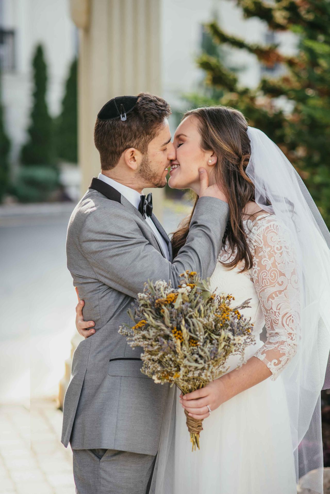 A bride and groom in wedding attire share a close embrace and kiss, with the bride’s veil and bouquet in the foreground.