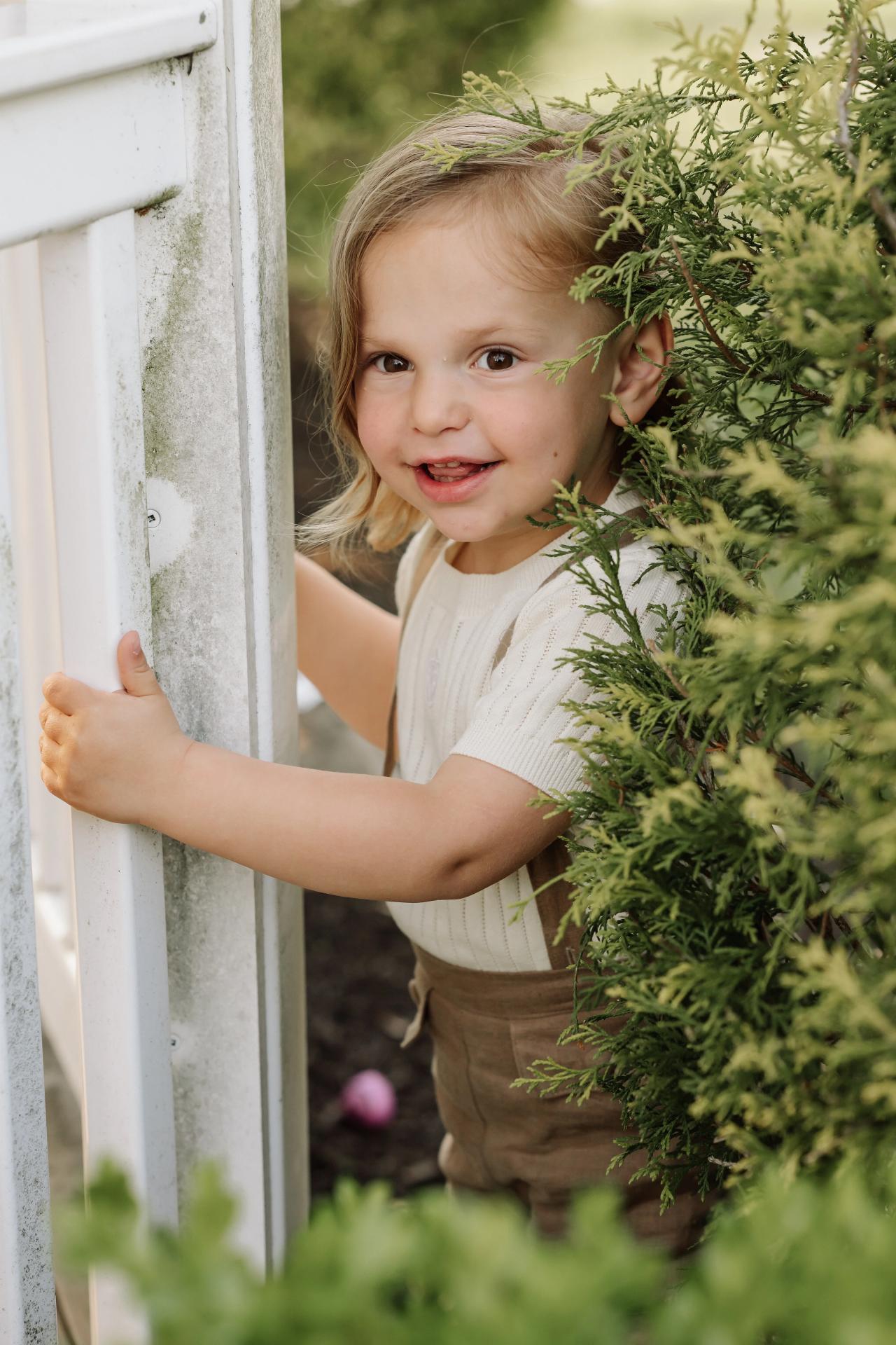 Child peeking through a white gazebo frame beside green shrubs