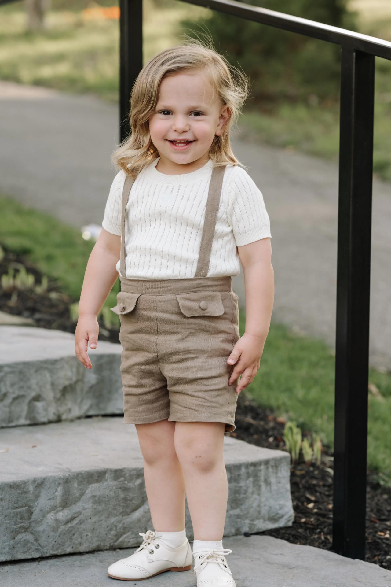 Smiling child with long hair and tan suspenders standing on stone steps