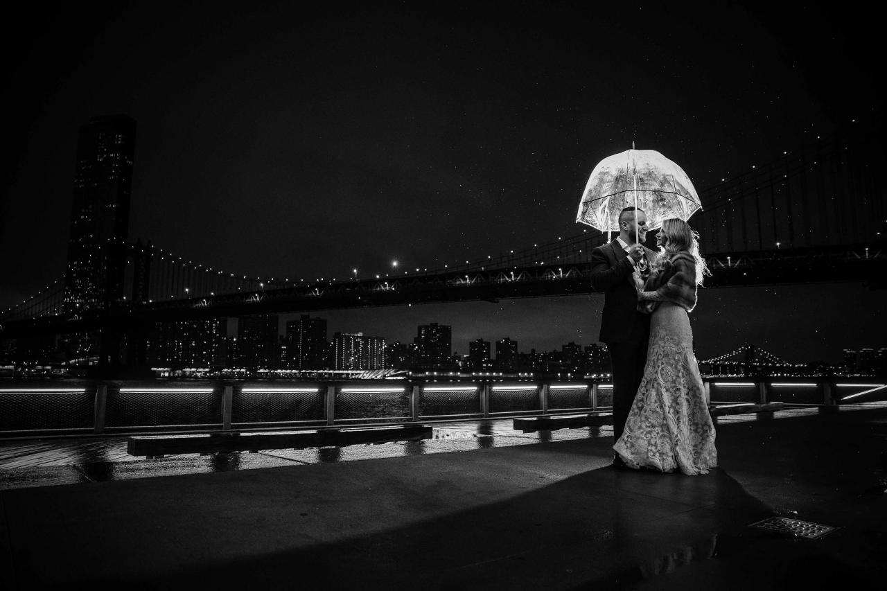 Black-and-white nighttime wedding photo of a bride and groom hugging under an umbrella on a waterfront bridge, with city lights in the background and light rain/droplets visible.