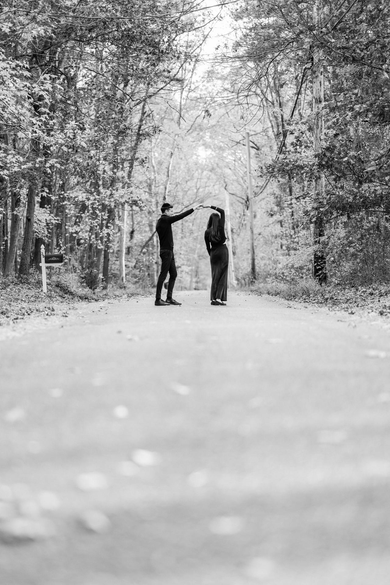 Black-and-white portrait of a couple twirling on a tree-lined road