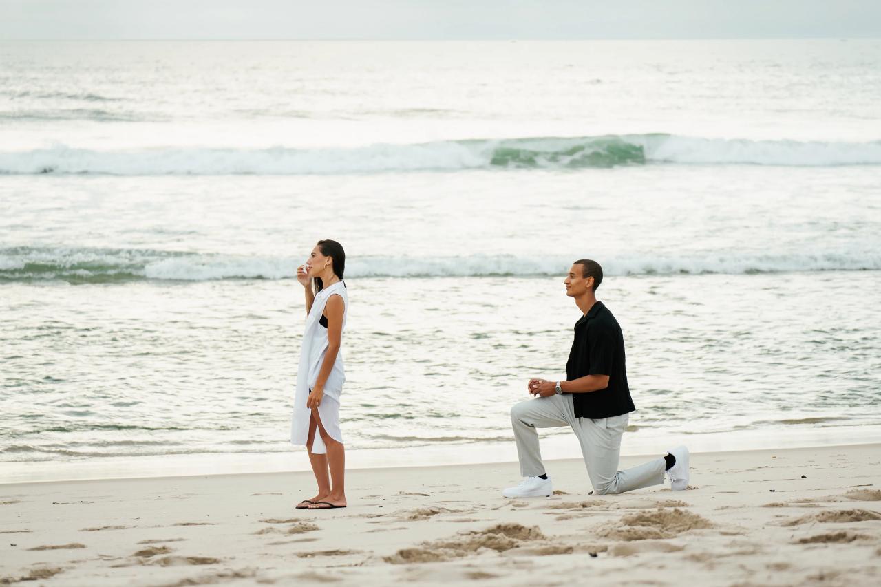 Man getting down on one knee behind his partner for a surprise beach proposal