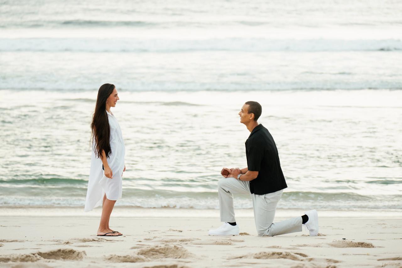Beach proposal with a man on one knee and ocean waves behind the couple