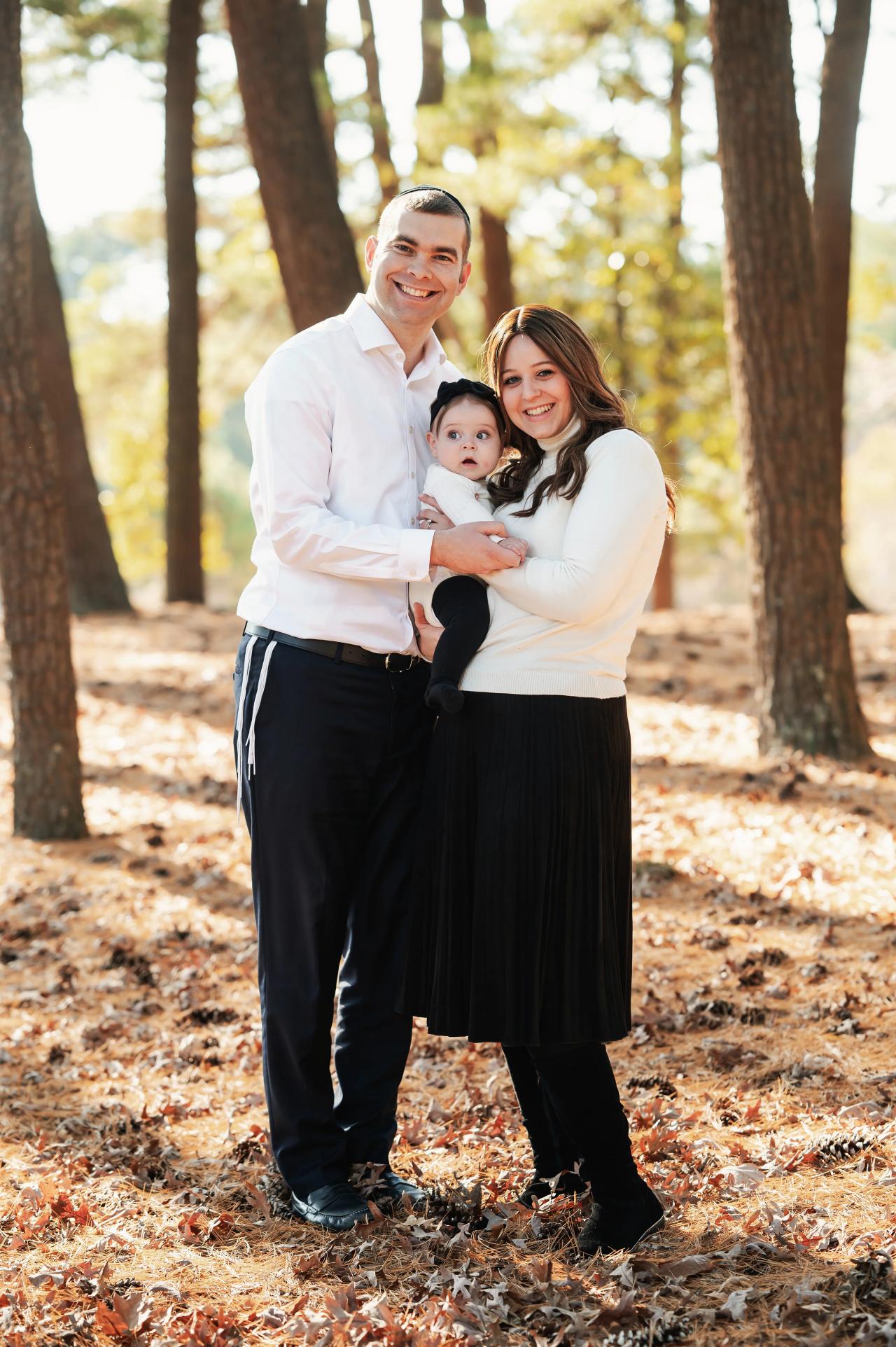 Family of three standing beneath tall trees during a fall photoshoot