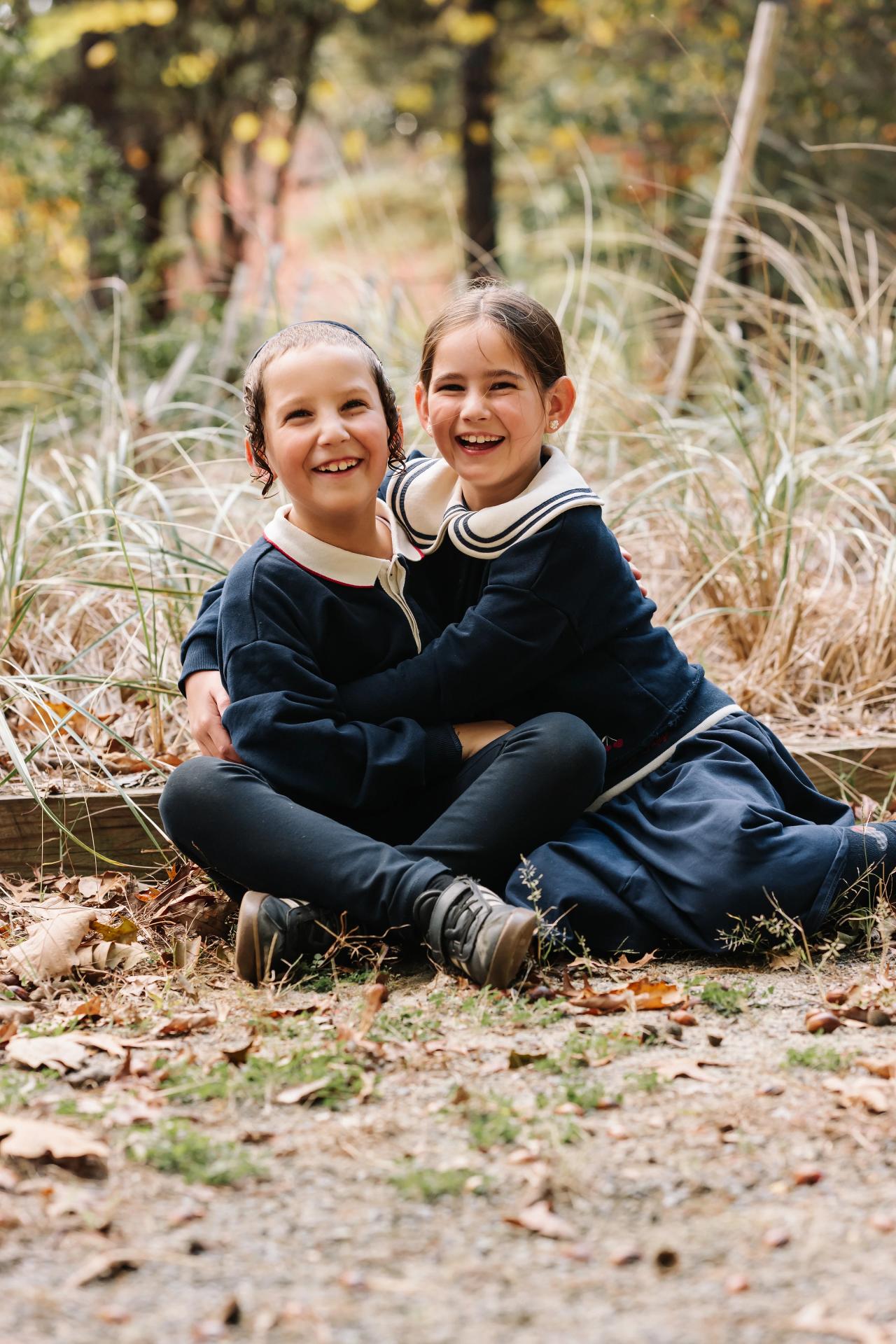 Brother and sister smiling together on a leaf-covered woodland path