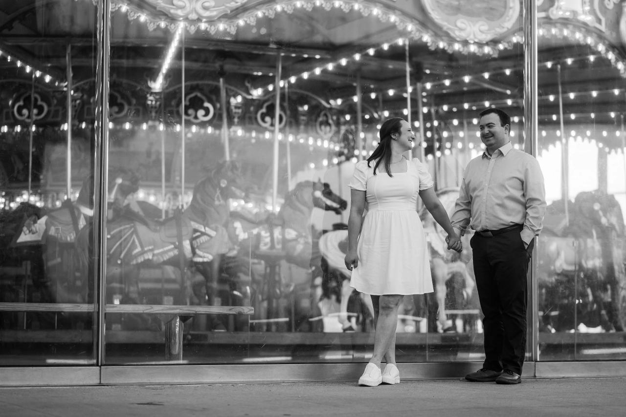 Black-and-white engagement portrait beside an illuminated carousel