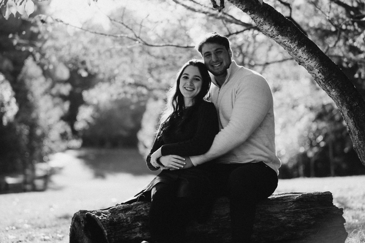 Black-and-white engagement portrait of a couple seated on a woodland log