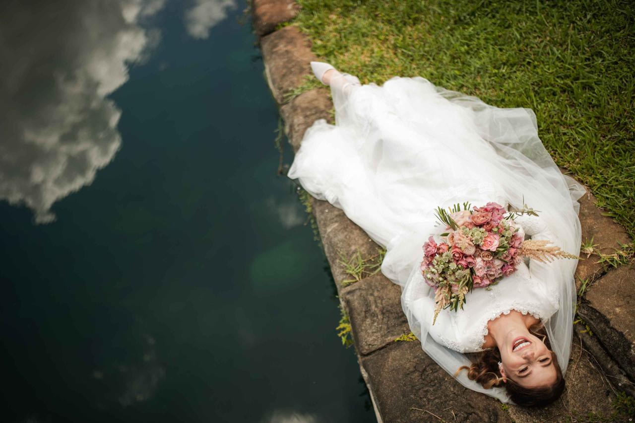 Bride in a white wedding veil reclining beside a pond, smiling with her bouquet resting in her hands on the stone edge.