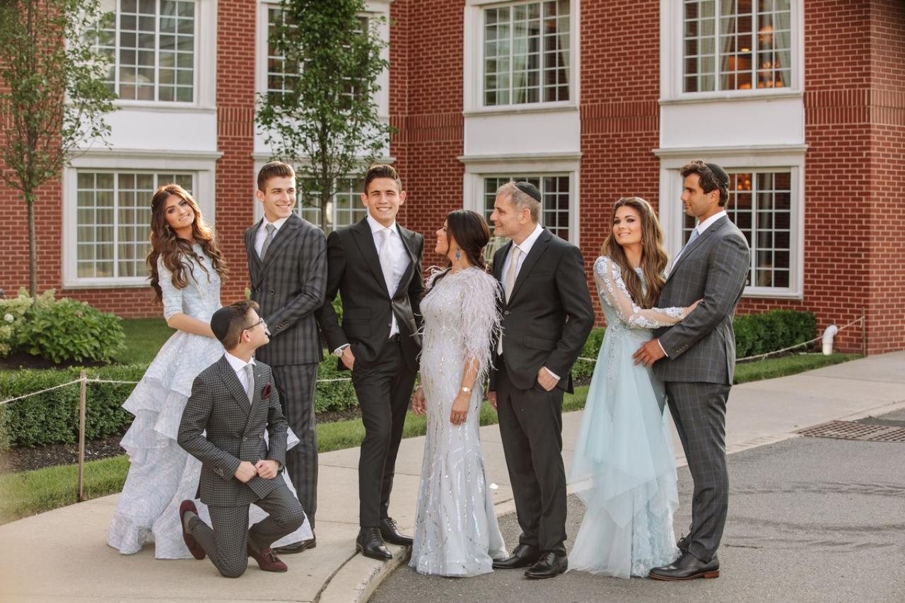 Wedding party outdoors in front of a brick building with windows: the groom kneels while bride and groomsmen and family members stand and face each other in a posed moment.
