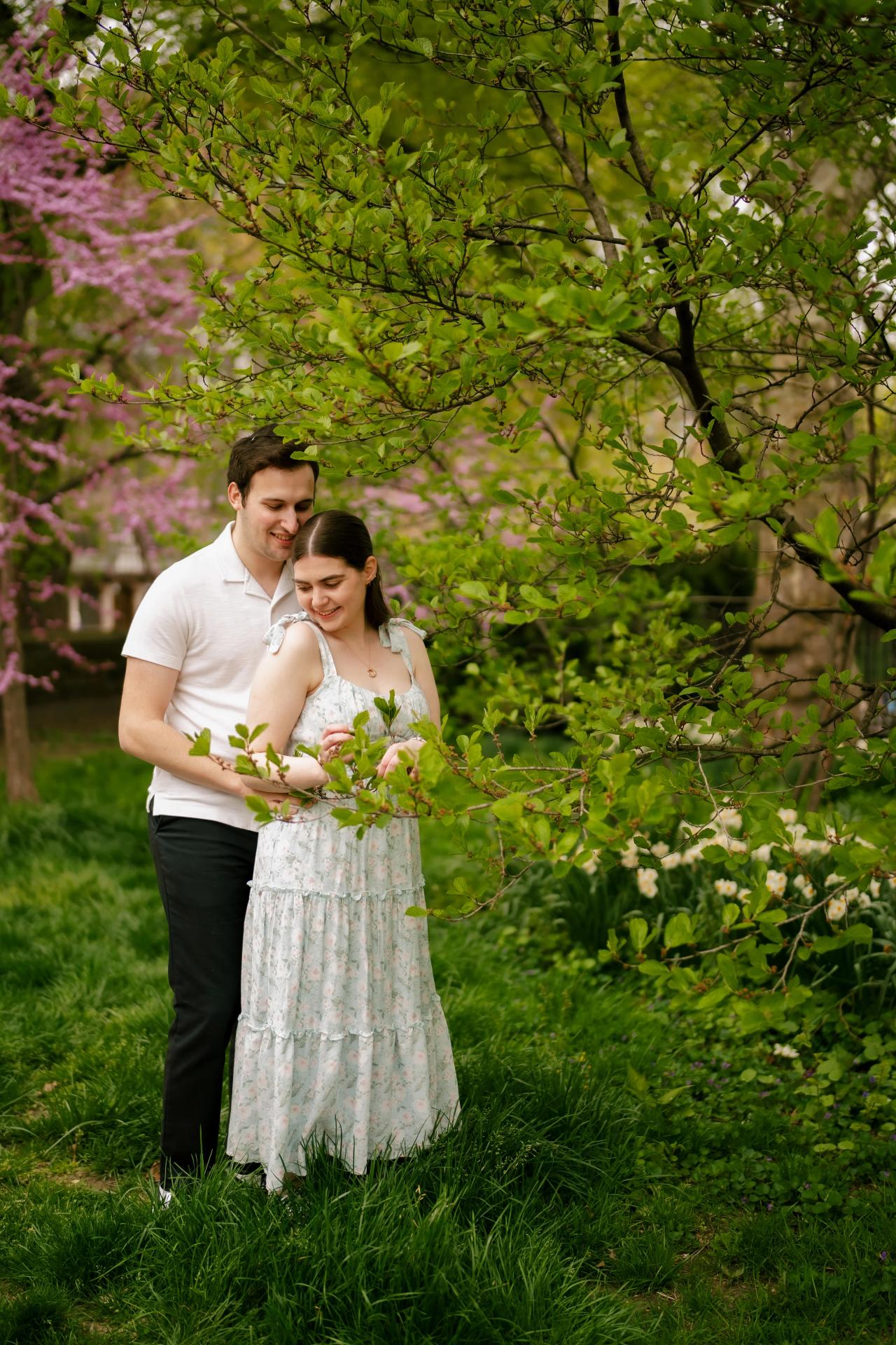 Full-length engagement portrait beneath green leaves and flowering branches