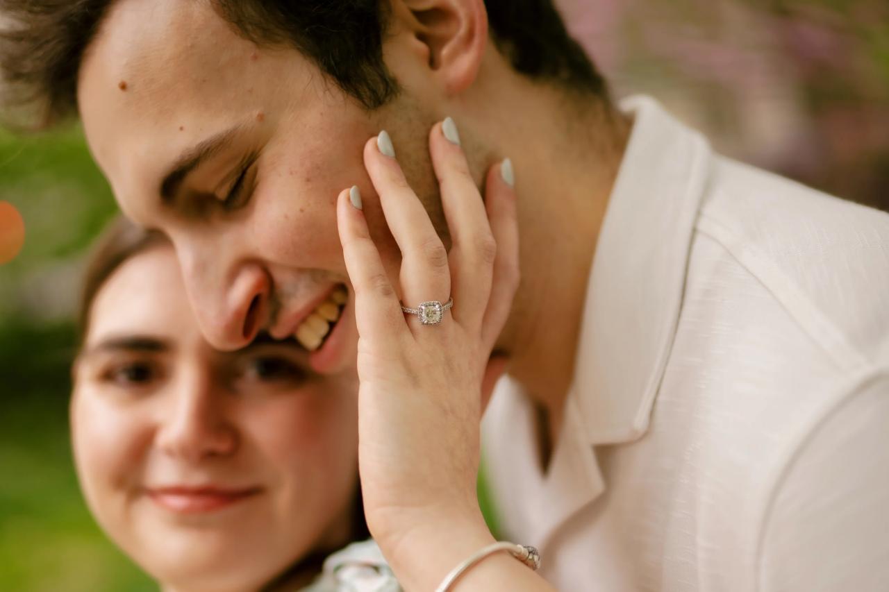 Close engagement portrait showing a hand and engagement ring against a partner’s cheek